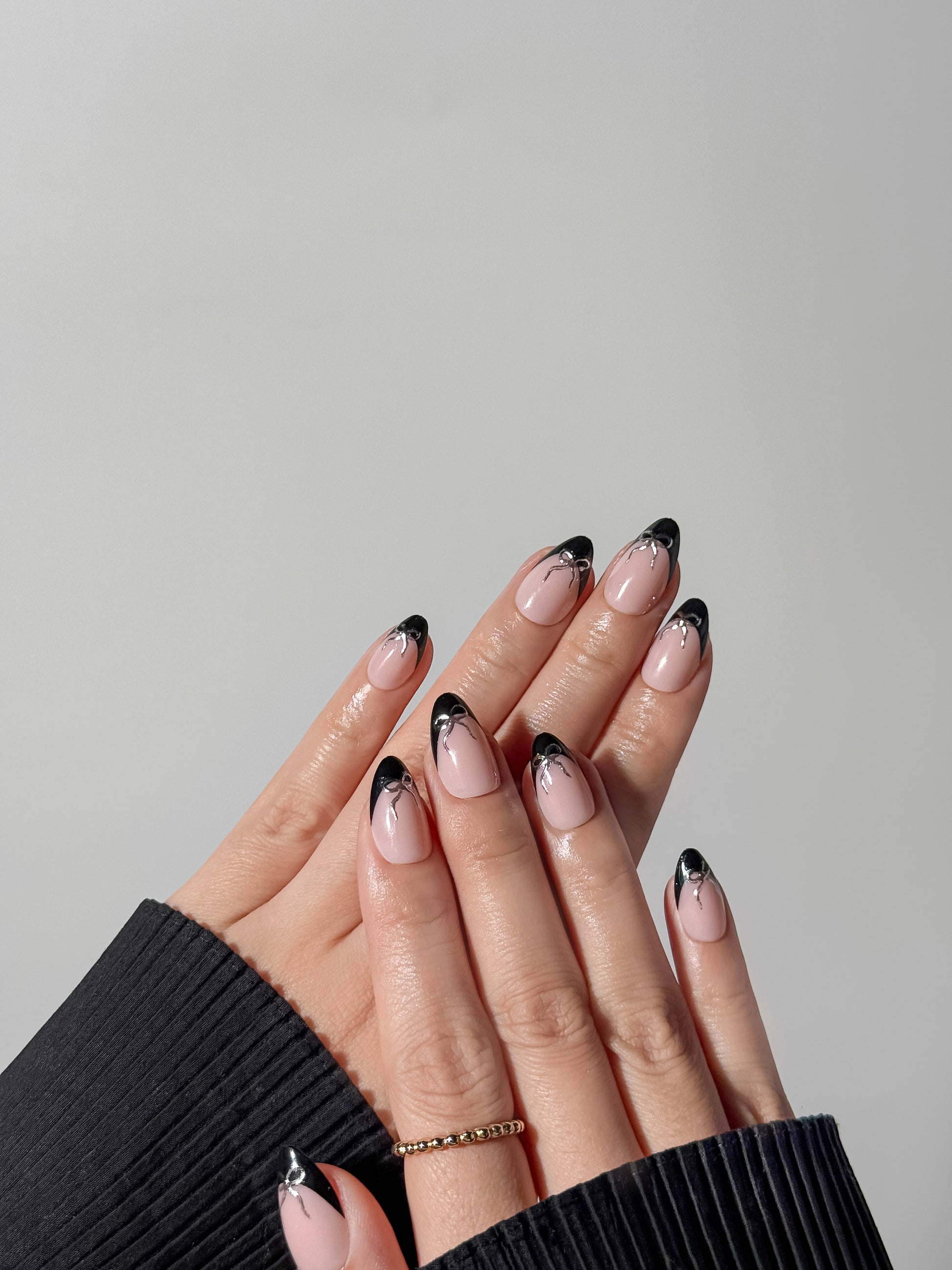 Model wearing almond-shaped black French tip nails with silver ribbon art, styled with gold rings