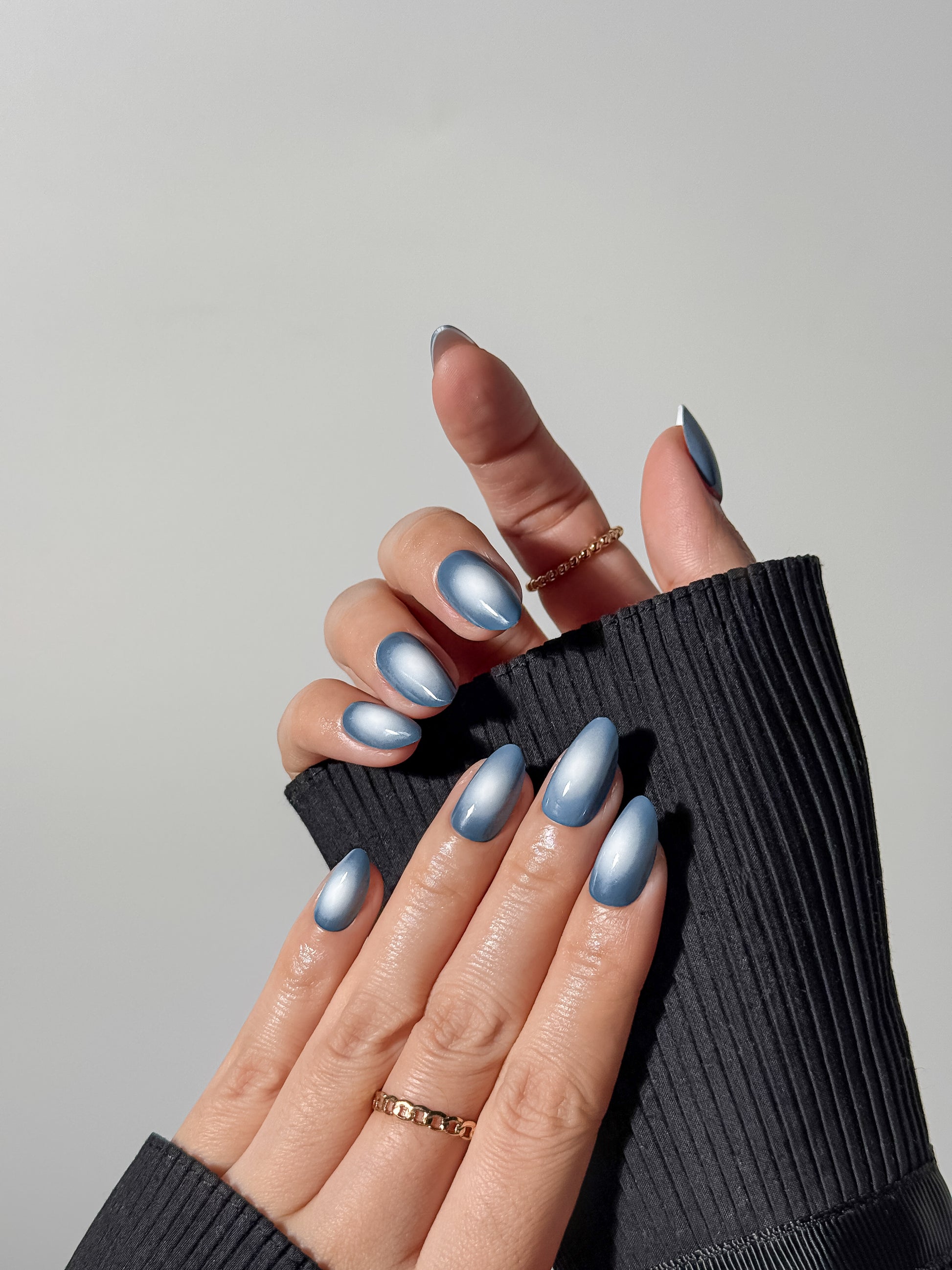 Hand wearing long almond blue velvet aura press-on nails styled with gold chain rings