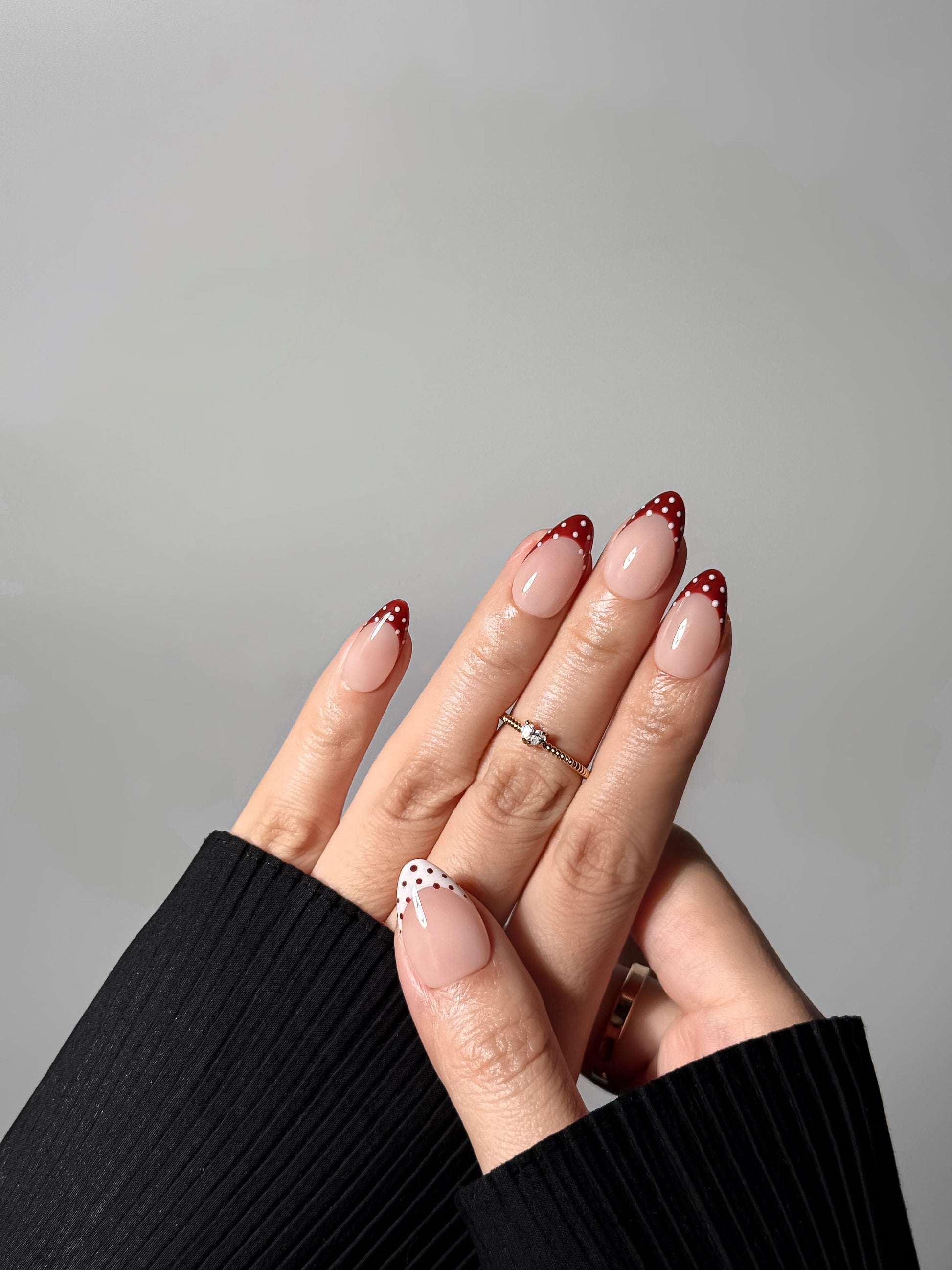 Elegant hand wearing almond-shaped nails with red and white polka dot French tips, accented by a small diamond ring.