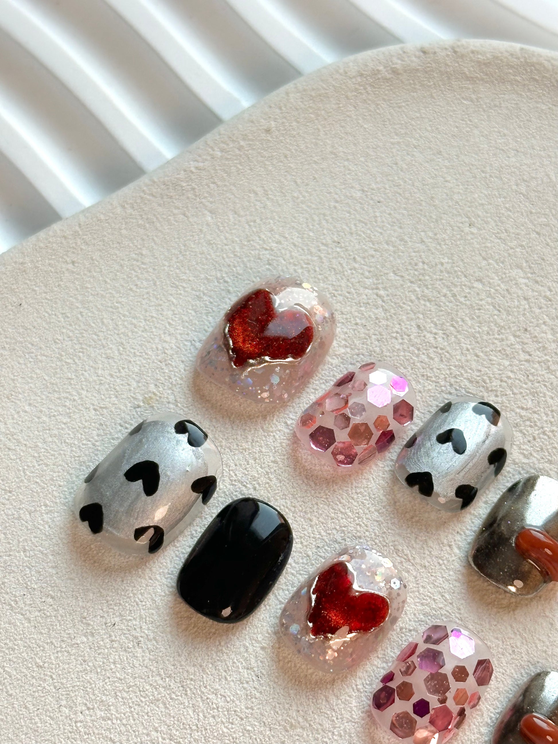 Extreme close-up of a press-on nail with a large shimmering red heart surrounded by iridescent glitter and chunky pink hexagonal sequins.