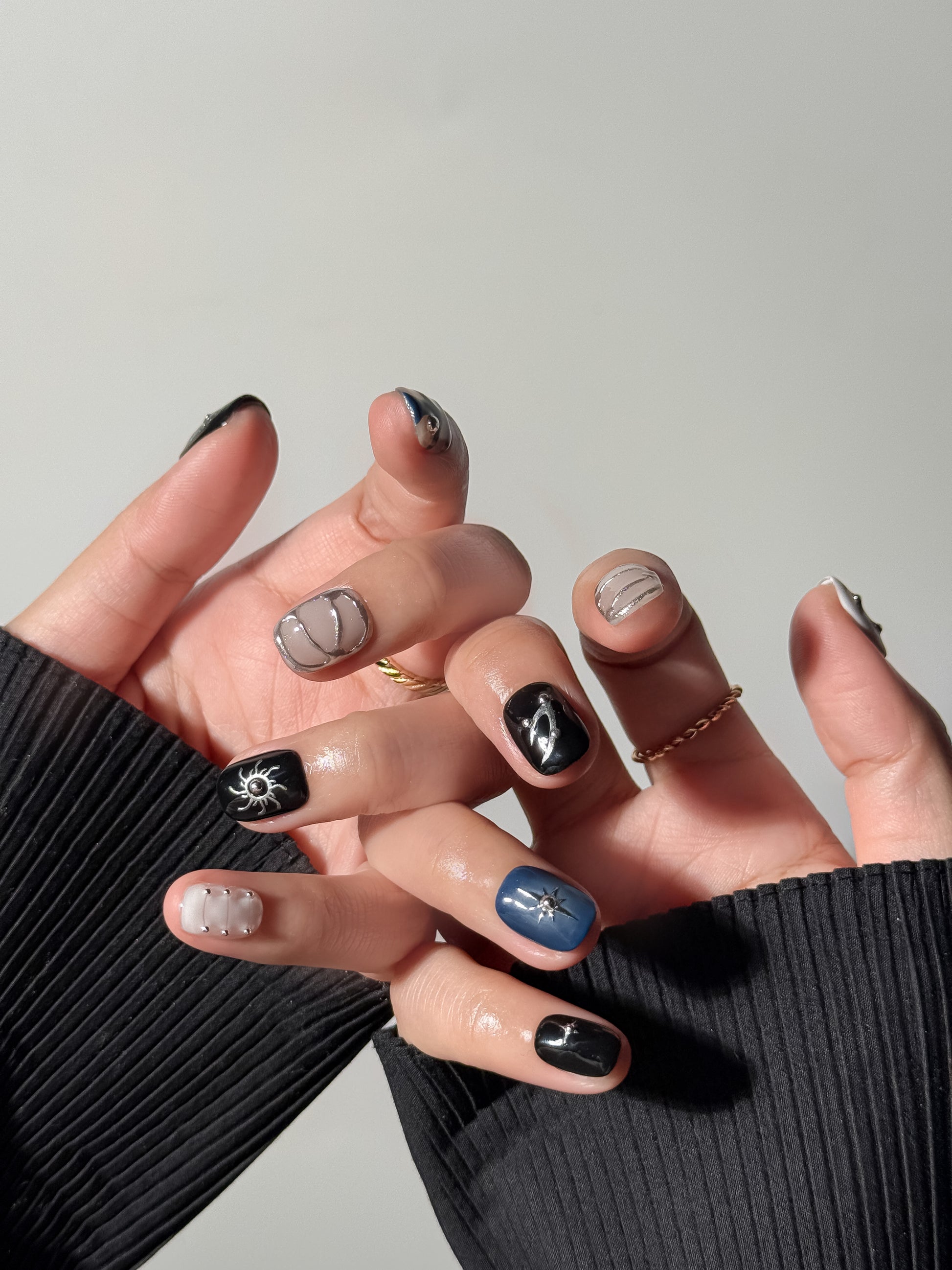 Both hands displaying a dark, celestial nail art design. The look features a mix of matte and glossy finishes, 3D silver star charms, metallic studs, and a deep blue accent nail with a silver North Star motif