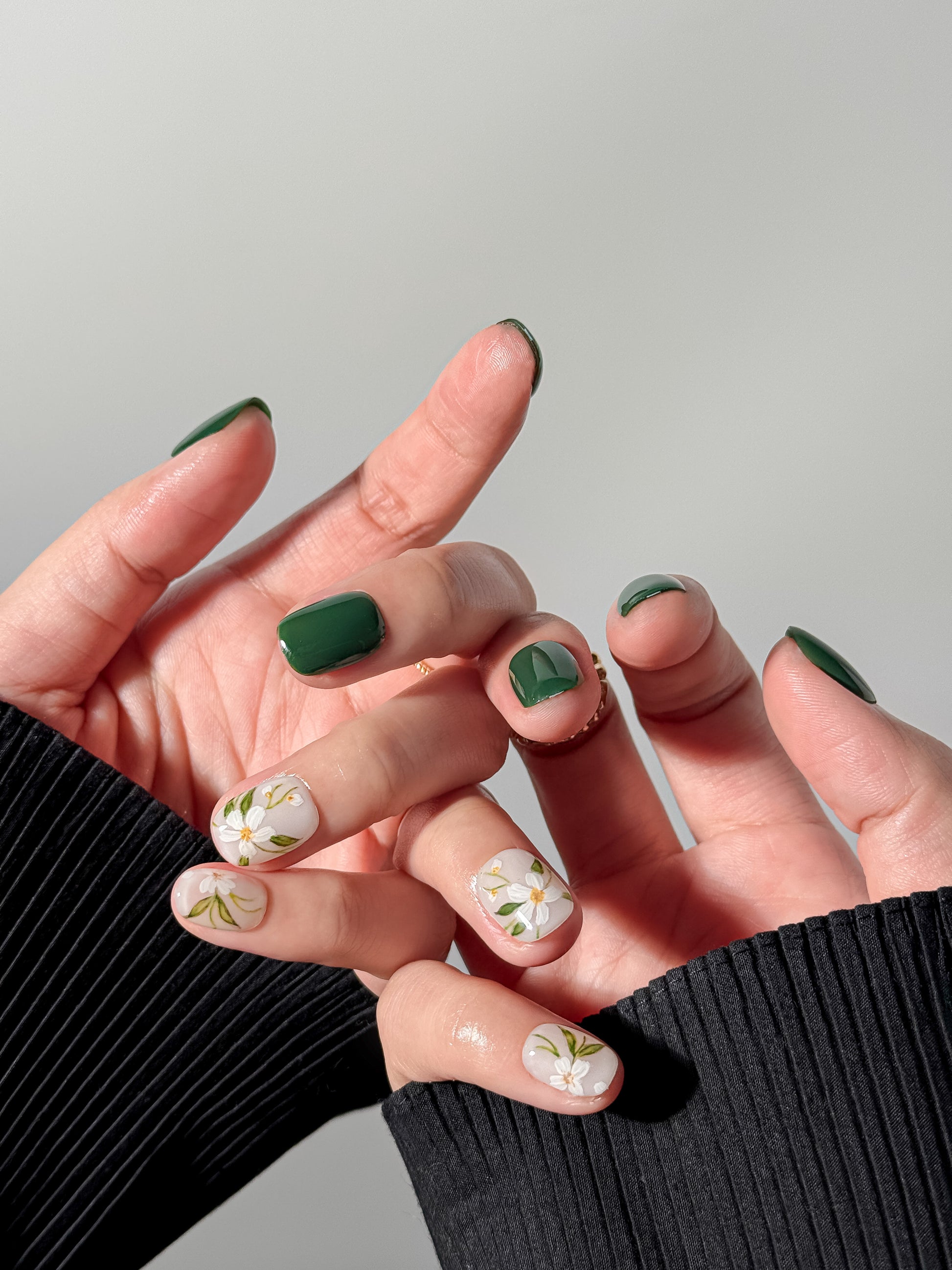 Two hands intertwined showcasing a sophisticated manicure with deep green gel polish and botanical floral art on the ring and pinky fingers, styled against a black ribbed sweater.