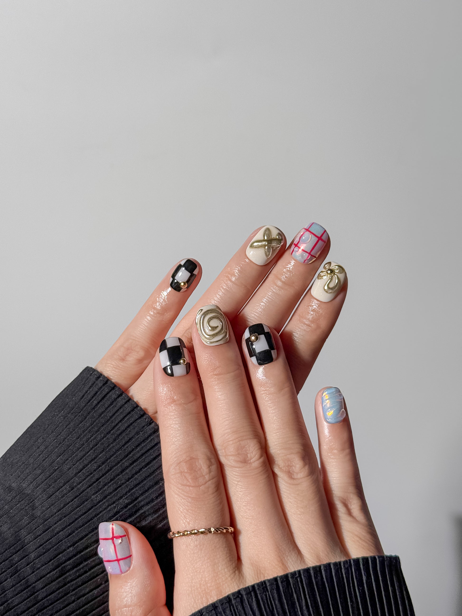 Two hands displayed against a neutral background showing a full eclectic manicure; the design includes 3D silver chrome textures, classic checkerboard nails with gold studs, and soft blue-pink iridescent aurora nails.
