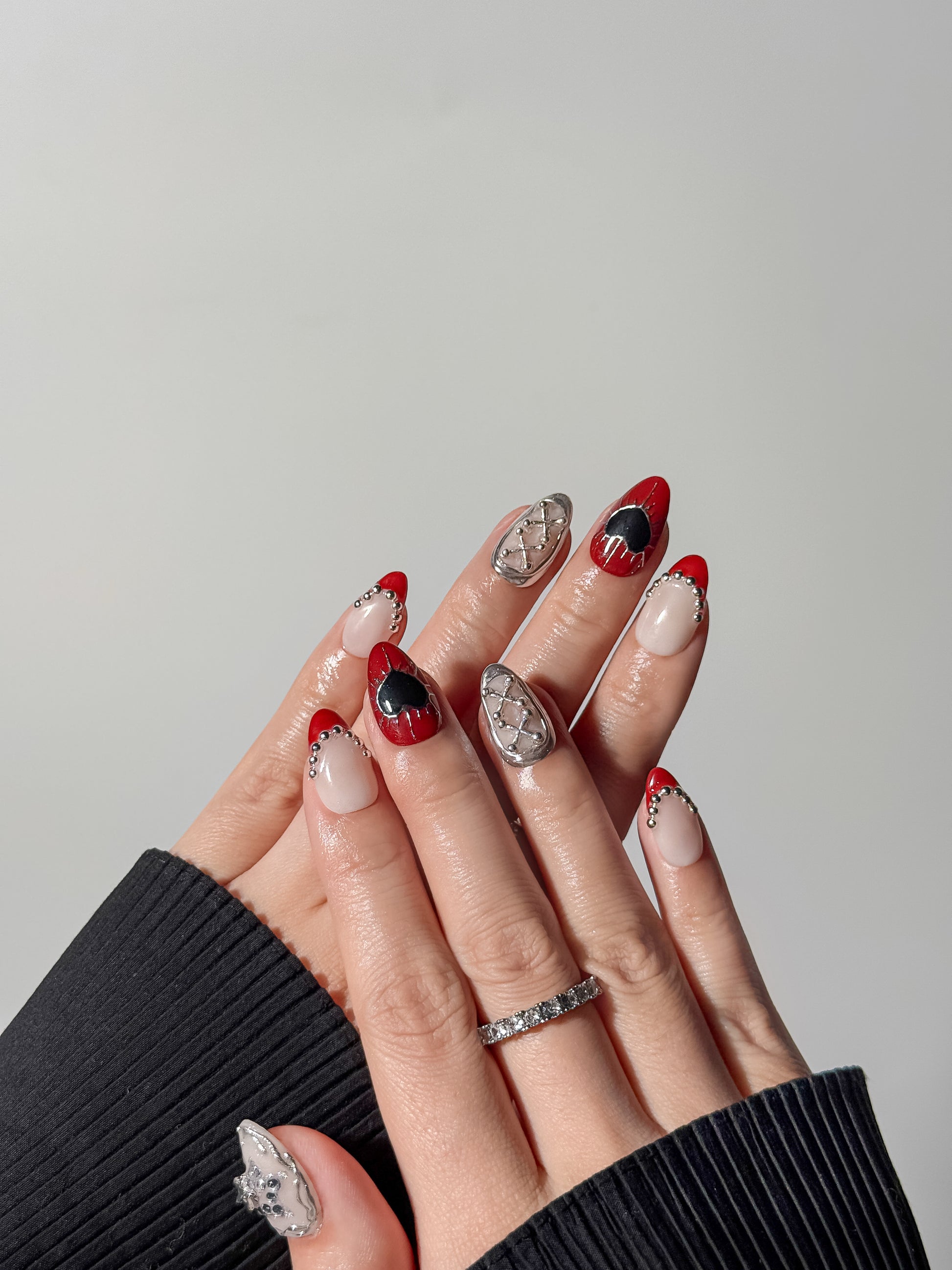 edgy almond nails in a grunge aesthetic style with silver 3d metallic textures and red and black heart designs on a hand with jewelry.
