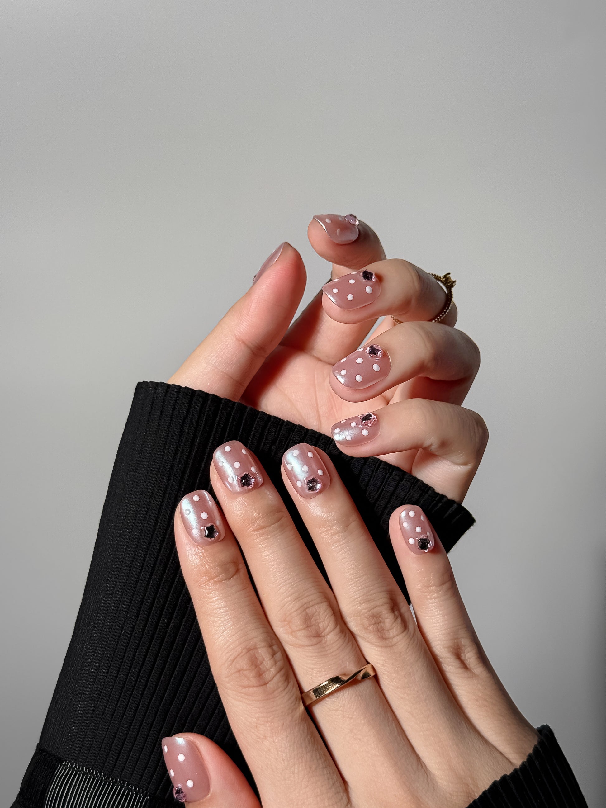 Two hands showing an elegant, shimmering pink manicure featuring a delicate white polka dot pattern and sparkling pink gems.