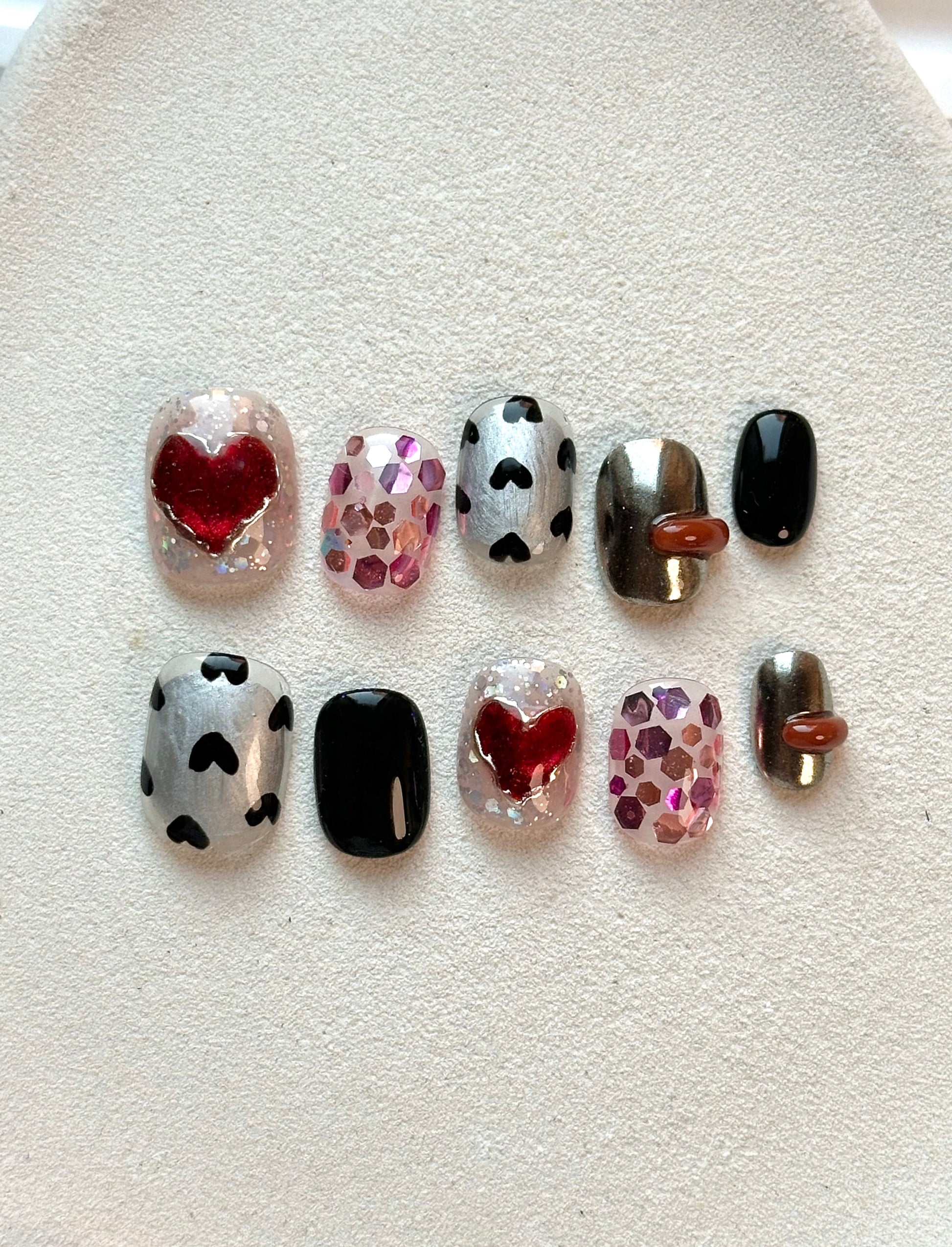 A flat-lay display of short, square press-on nails featuring metallic silver with black hearts, chunky pink glitter, and 3D red heart designs.