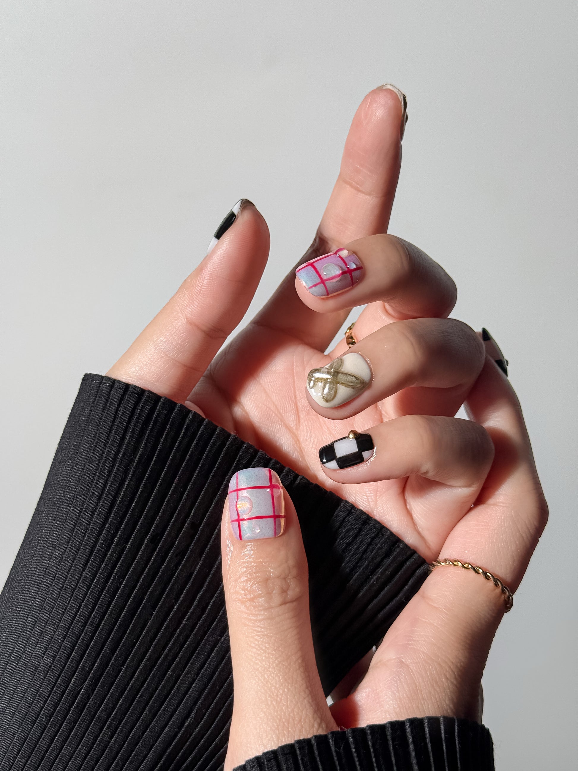 A close-up shot of a hand wearing short square nails; the thumb features a red grid plaid over an iridescent base with clear 3D gel droplets, paired with a black checkered index finger and gold ring.