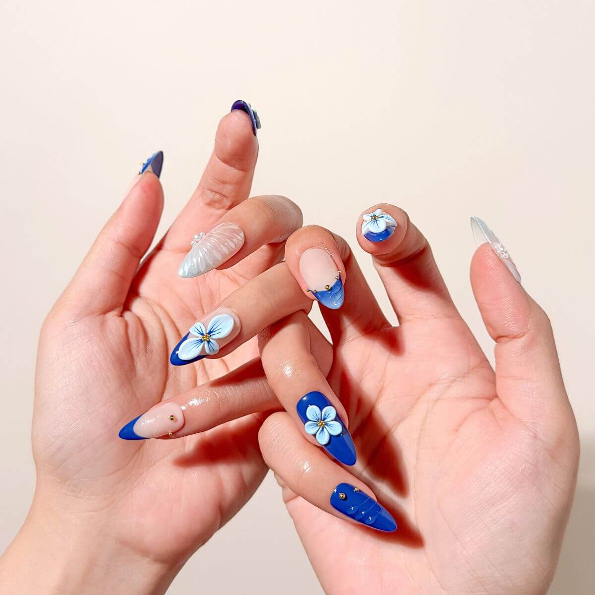 Elegantes Lifestyle-Foto von zwei ineinandergelegten Händen mit royalblauen künstlichen Fingernägeln, kunstvollen 3D-Blüten und schimmernden Mermaid-Effekten.