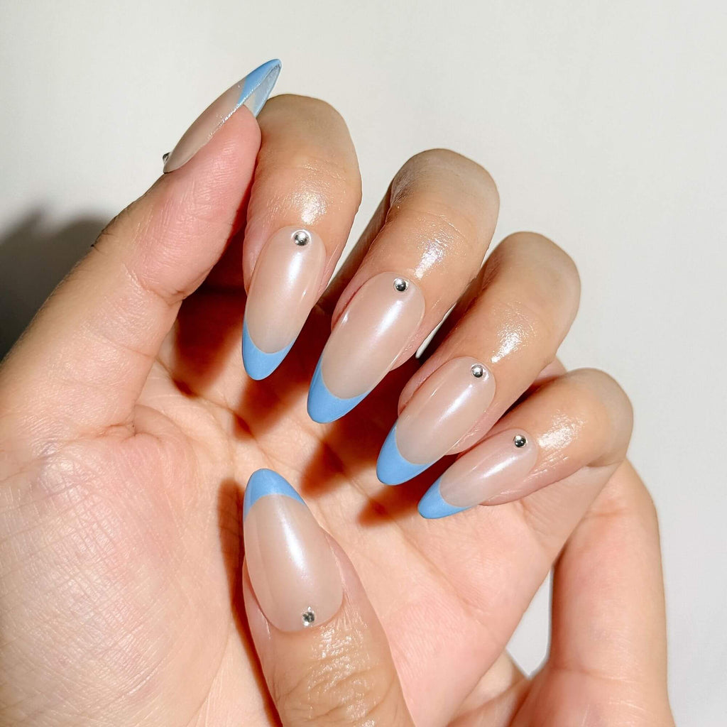 Detailaufnahme einer Hand, die den reflektierenden Chrome-Glanz auf handgefertigten blauen French-Nägeln sowie die präzise Mandelform und den gesetzten Strassstein zeigt.