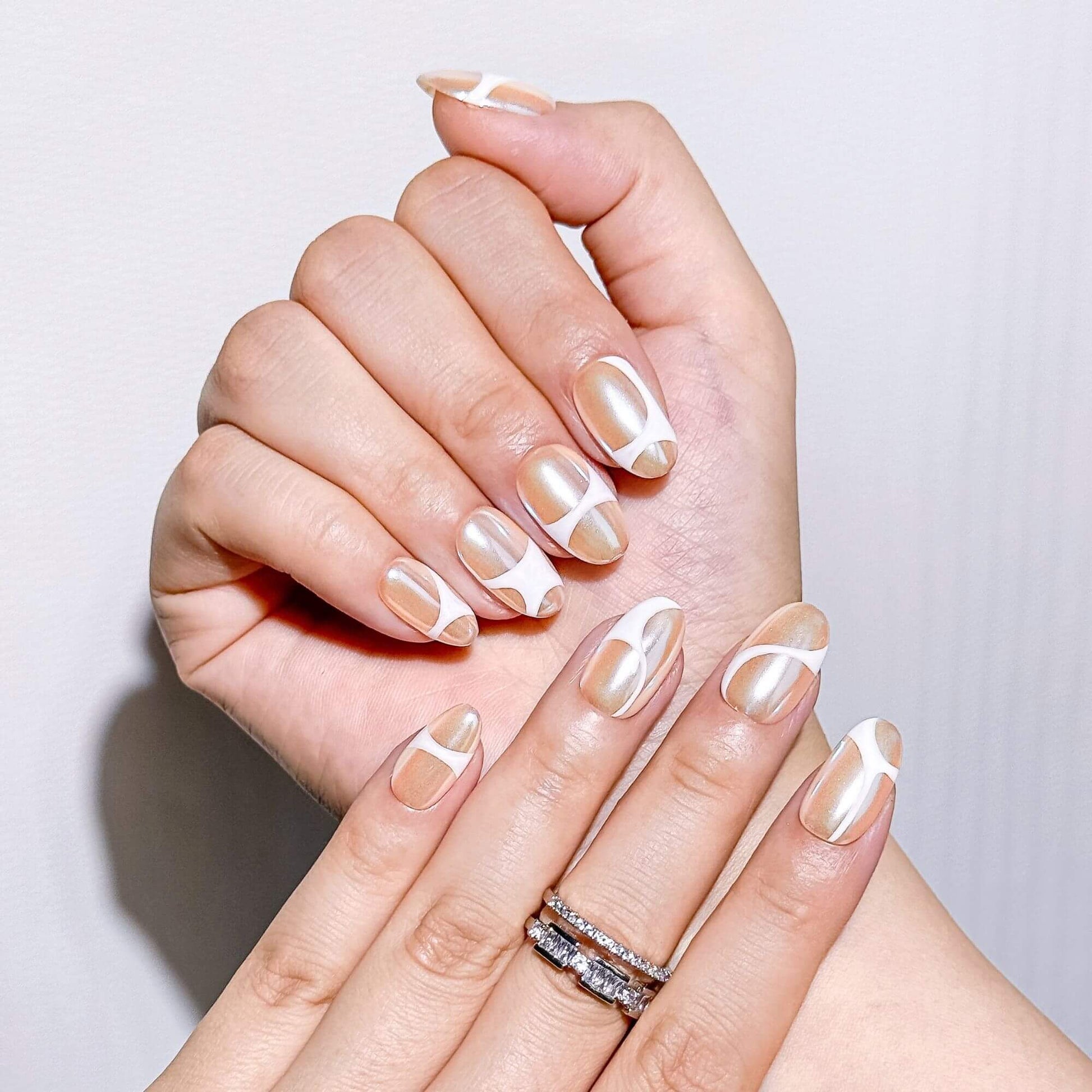 Nahaufnahme von künstlerisch handgefertigten Champagne Glaze Chrome-Nägeln mit weißem Liniendesign und silbernen Ringen.