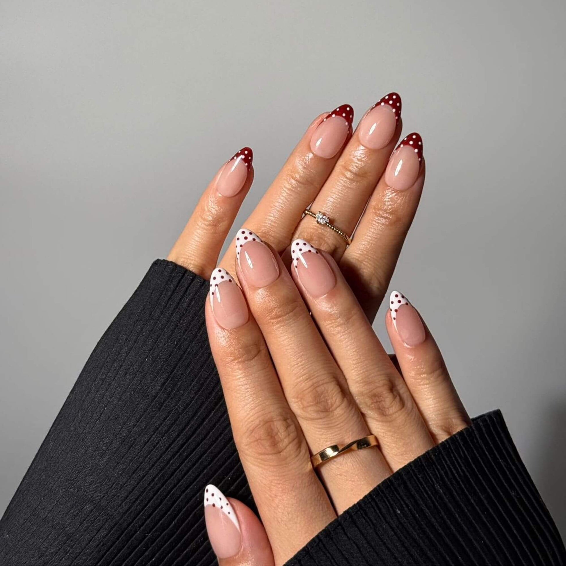 Ein professionelles Lifestyle-Foto von Händen mit Cherry Mix Kunstnägeln, die eine Kombination aus rot und weiß gepunkteten French-Tips auf einer mandelförmigen Nude-Basis zeigen.