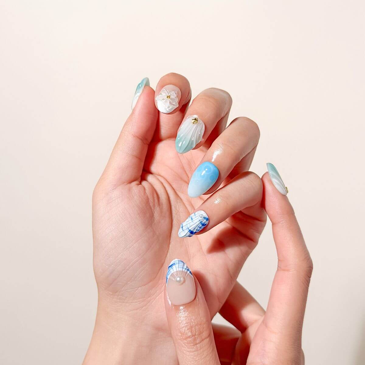 Close-up photo of a hand wearing seafoam blue artificial nails highlighting a textured shell pattern, a clear 3D flower blossom, and subtle blue plaid tips.