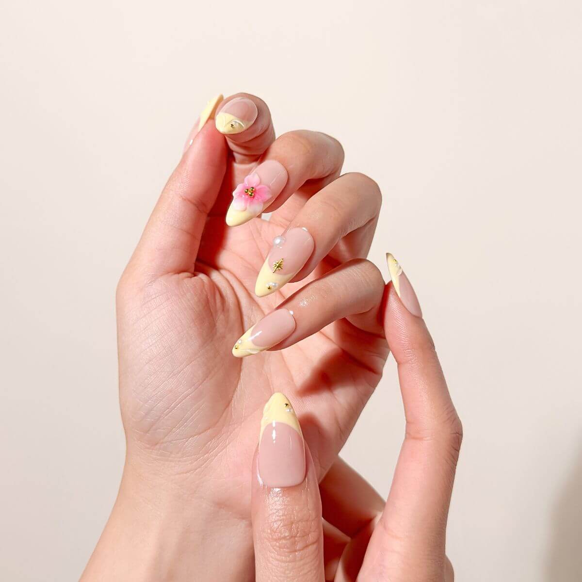 Nahaufnahme einer Hand mit pastellgelben French-Kunstnägeln, akzentuiert mit einer zartrosa 3D-Blüte und goldenen Sternen-Details.