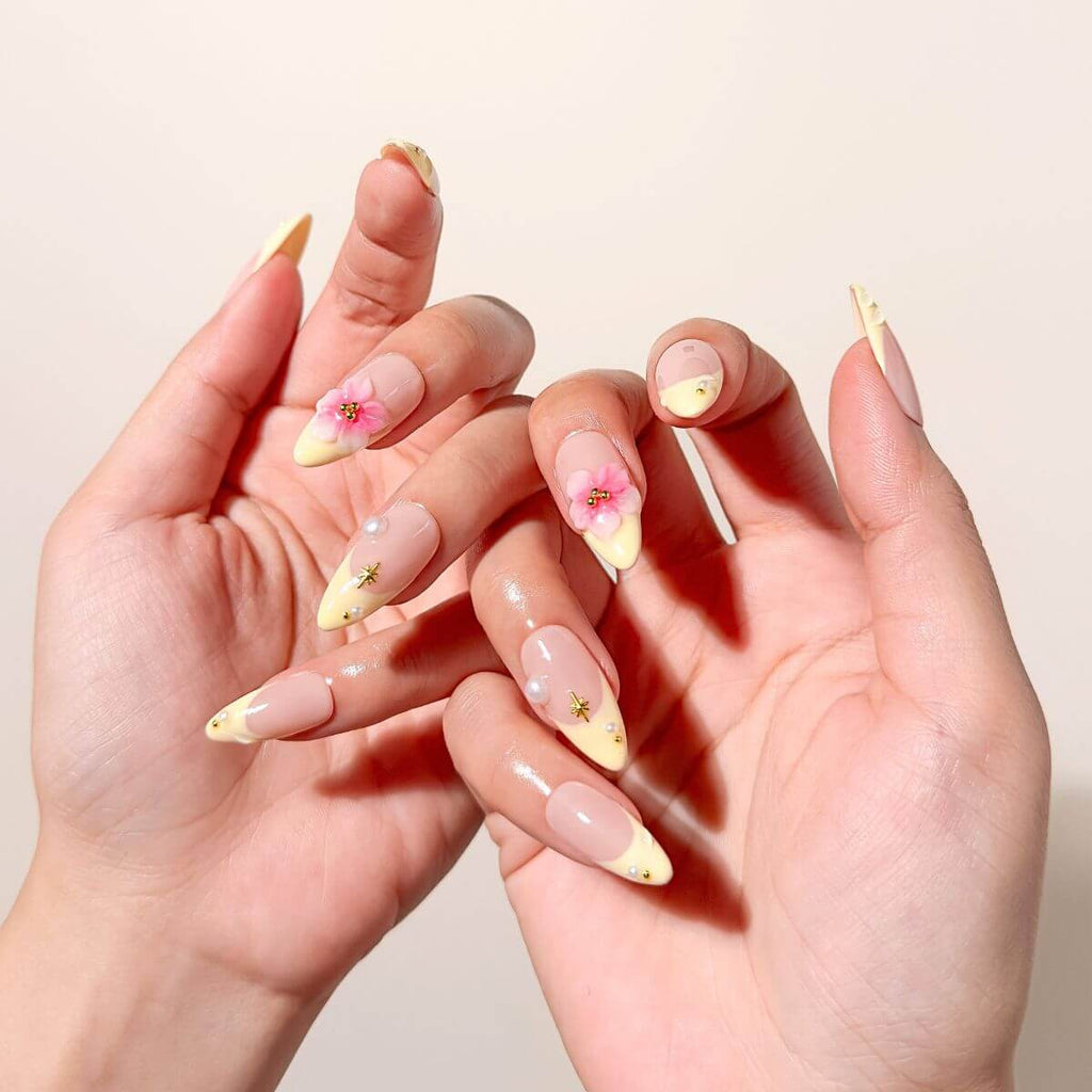 Lifestyle-Foto von zwei Händen mit buttergelben Kunstnägeln in Mandelform, verziert mit dreidimensionalen Blumen im Coquette-Stil und kleinen Perlen.