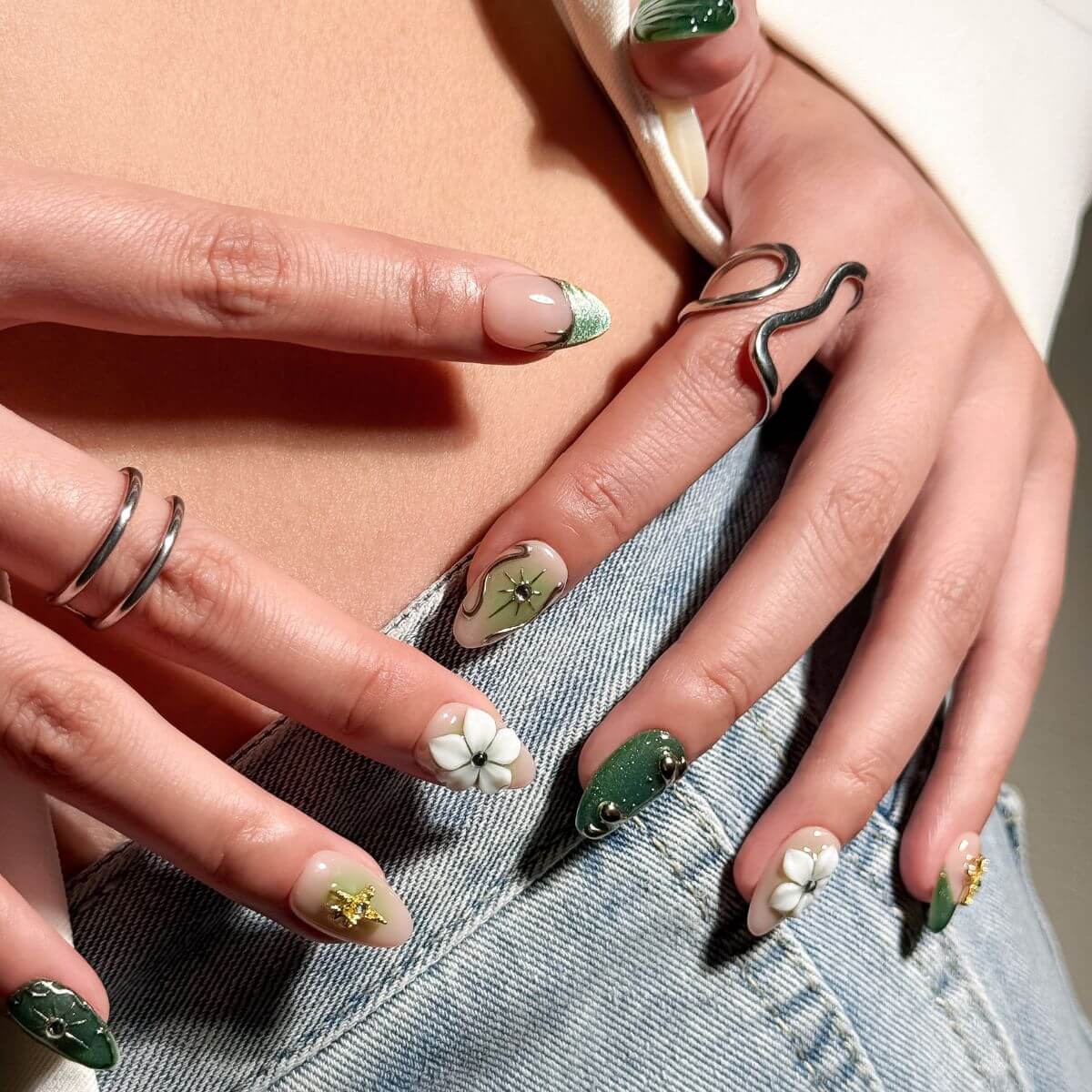 Ein Lifestyle-Foto von künstlichen Fingernägeln zum Aufkleben in Grün mit Cat-Eye-Effekt und metallischen Y2K-Verzierungen, fotografiert vor einer Jeans.