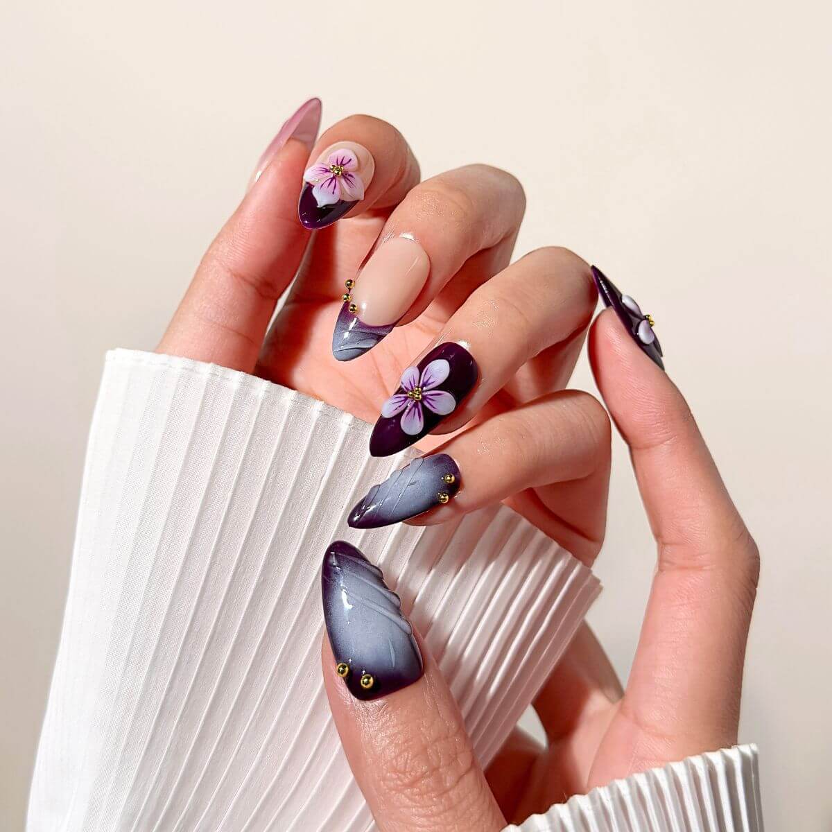 Close-up of a hand wearing dark purple almond-shaped glue-on nails with intricate 3D floral accents, a smoky grey magnetic finish, and small gold beads.