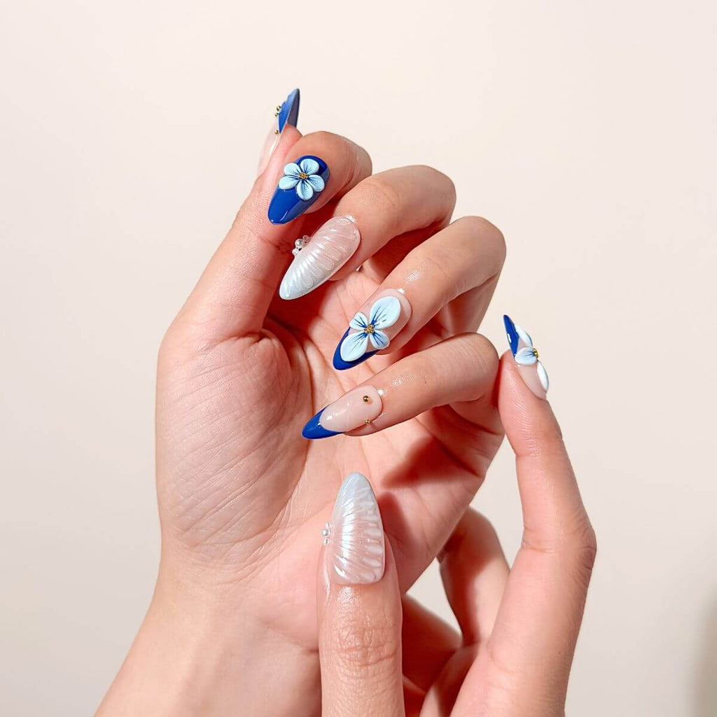 Close-up of a hand wearing cobalt blue almond-shaped glue-on nails featuring textured pearl seashell art, hand-painted 3D flowers, and classic French tips with gold studs.