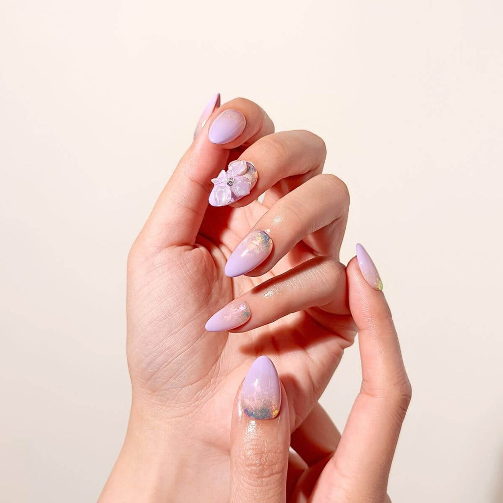 Elegant close-up of a hand showcasing a pastel lilac almond press-on manicure under bright lighting, focusing on the textured gel flower petals and reflective glass-foil details.