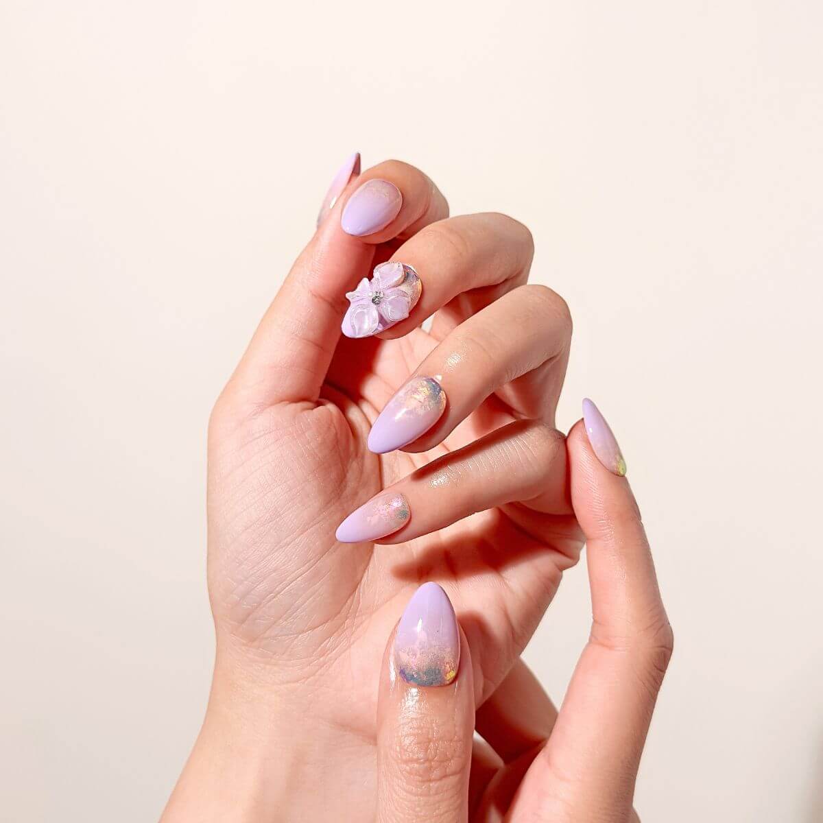 Elegant close-up of a hand showcasing a pastel lilac almond press-on manicure under bright lighting, focusing on the textured gel flower petals and reflective glass-foil details.
