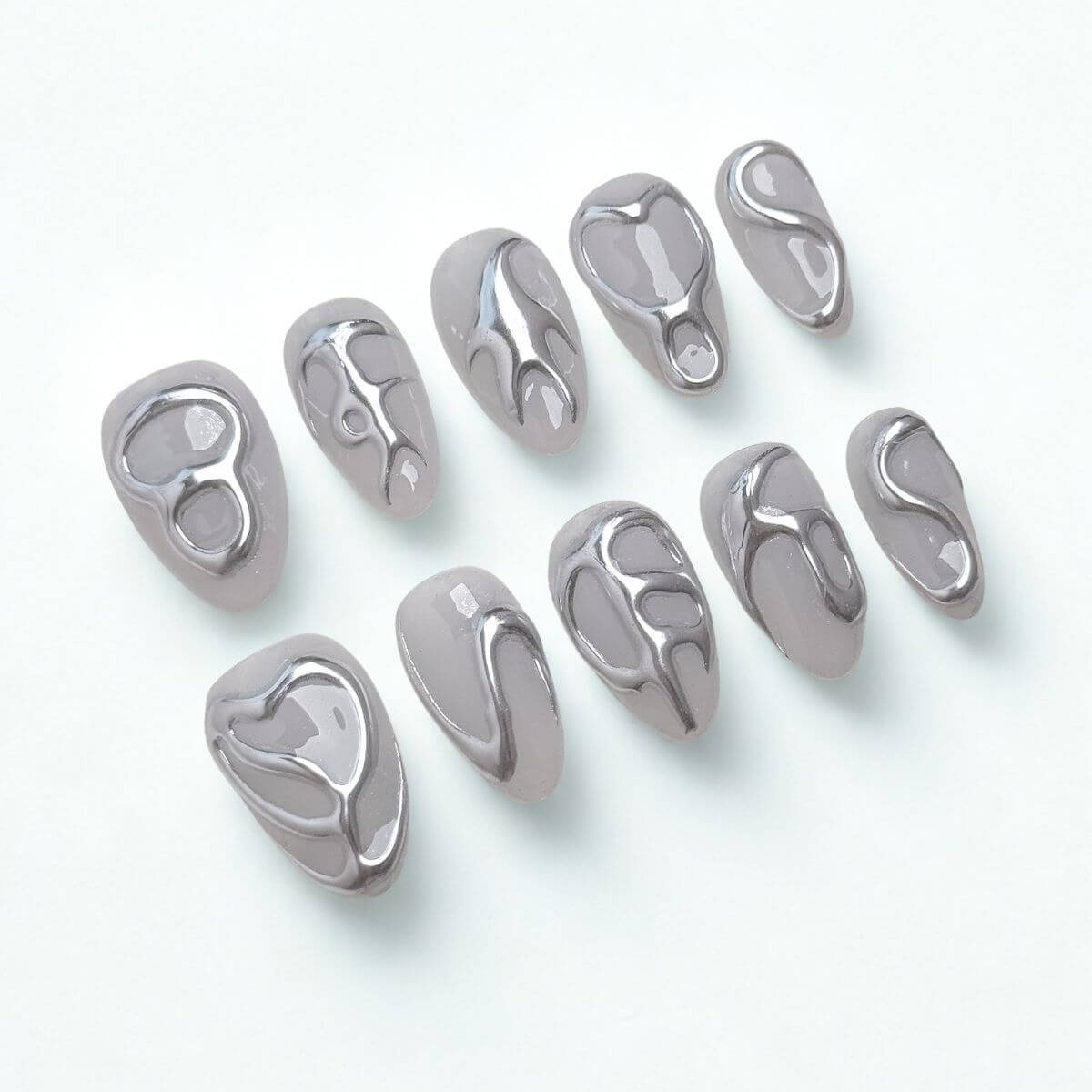 Flatlay von 10 justmee Kunstnägeln in Mandelform. Milching-graues Gel-Design mit 3D Chrome Liquid Metal Effekten für eine moderne Maniküre zum Selbermachen.