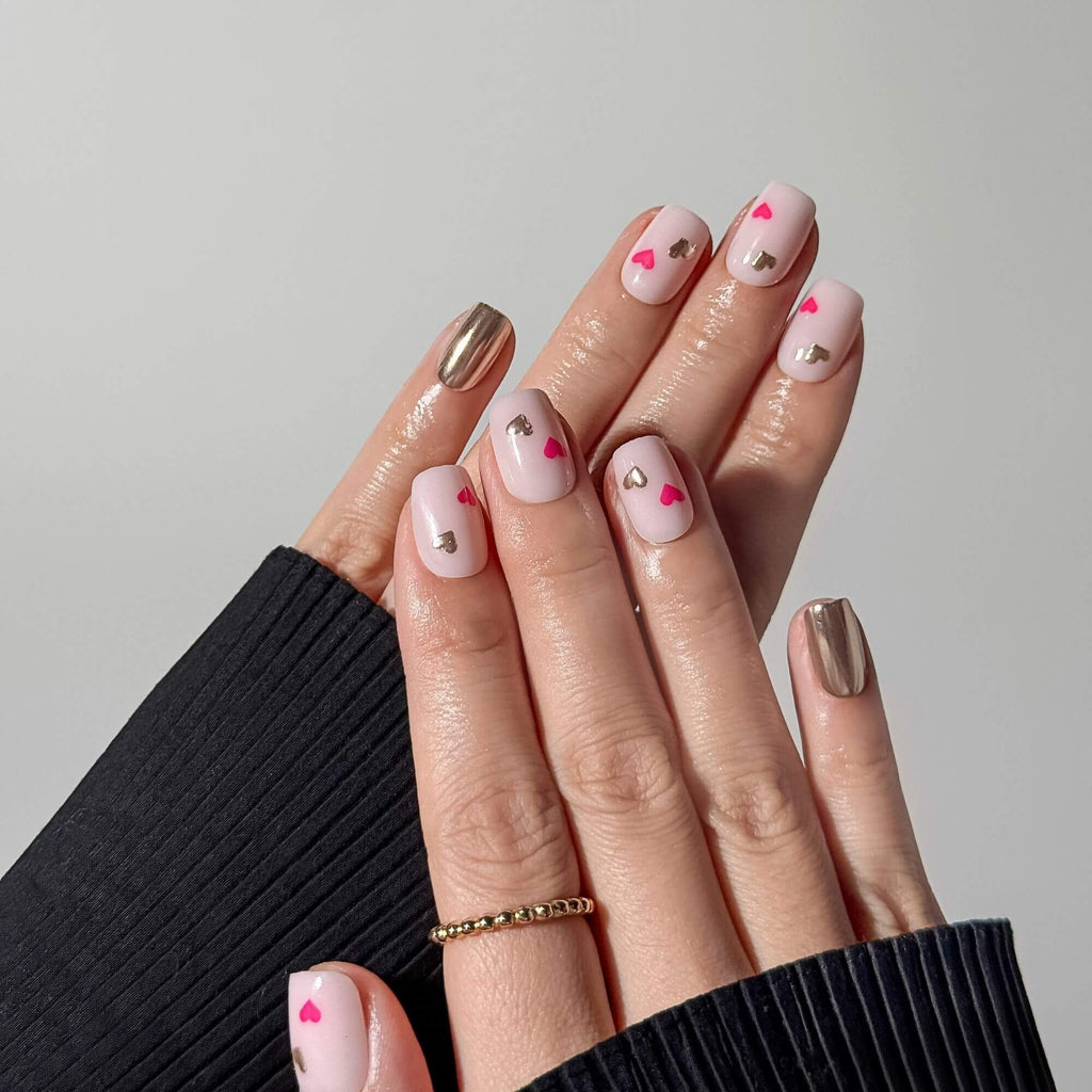 Zwei Hände, die ein komplettes Set kurzer eckiger Press-on-Nägel in Milchrosa mit silbernen Chrom-Akzenten und Herzmustern für einen modernen, romantischen Look präsentieren.