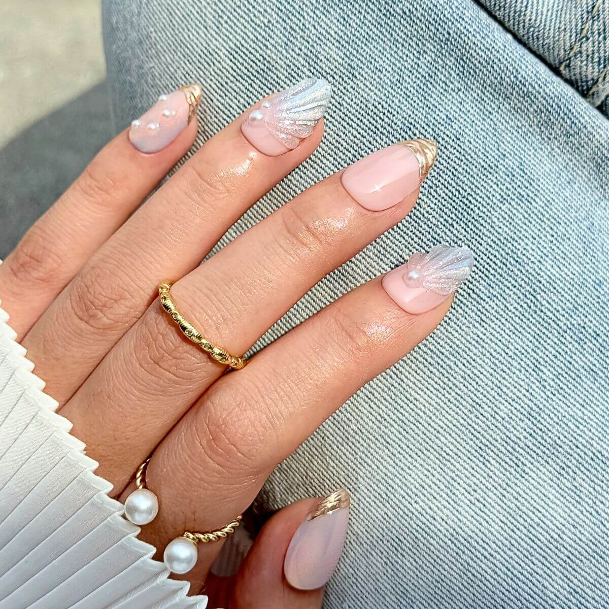 Close-up of a hand wearing pearlescent 3D shell press-on nails with liquid-gold french tips in outdoor daylight, styled with pearl-embellished rings and light blue denim.