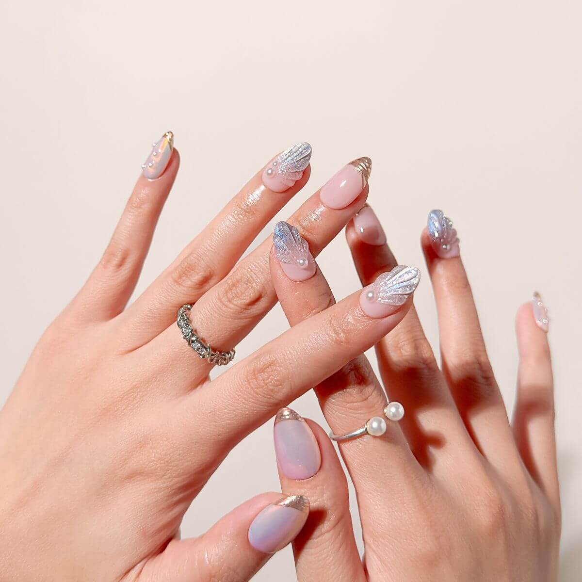 Verschränkte Hände eines Modells mit minimalistischen Perlenringen präsentieren die Pearl Siren Maniküre und zeigen den dynamischen rosafarbenen Multichrome-Schimmer und die strukturierte Muschelkunst.