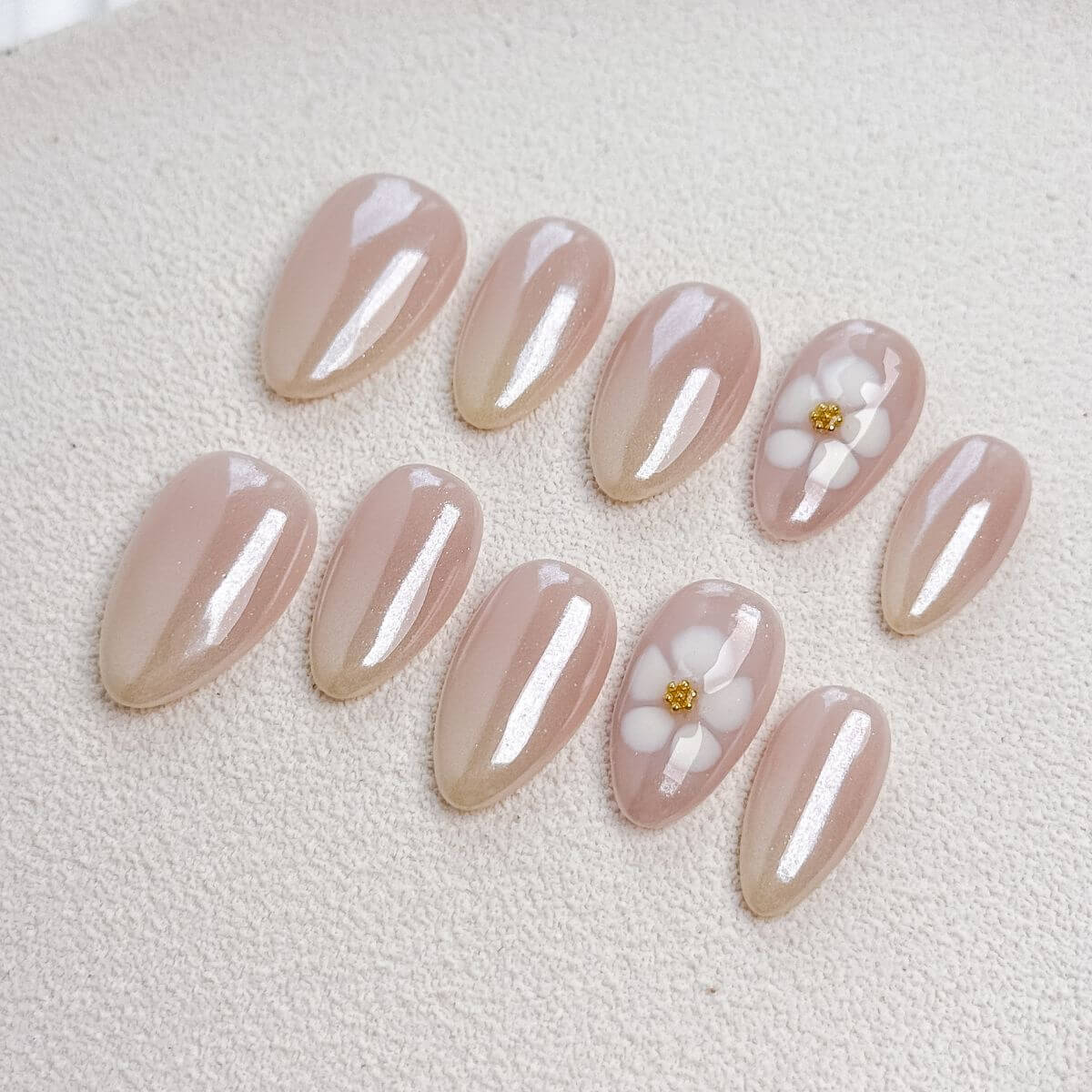 Studio-Draufsicht auf luxuriöse Press-On Nägel in mittlerer Mandelform mit zartem, perlmuttglänzendem Chrome-Nude-Base und plastisch modellierten weißen Blüten mit goldenem Zentrum.