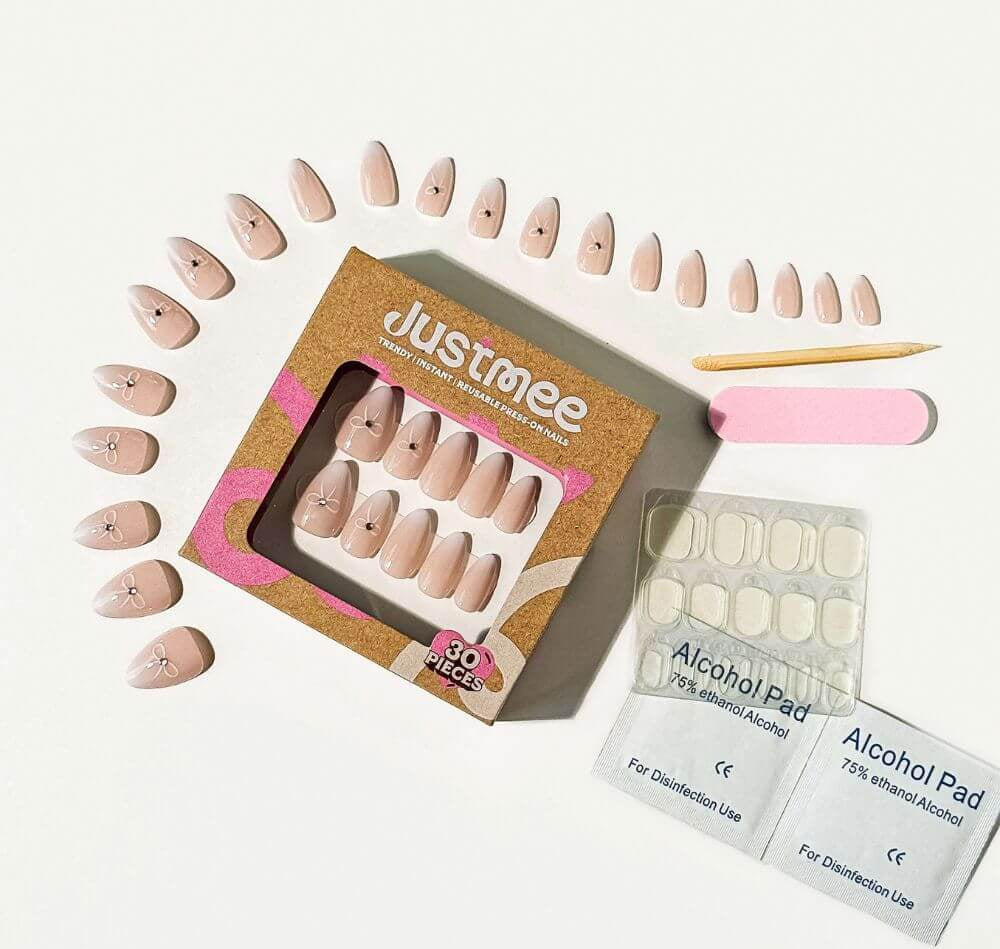 An overhead shot of a full "Justmee" press-on nail kit on a white surface, featuring an arched arrangement of individual nails, a cardboard box, and essential application tools like alcohol pads, an orange stick, and a nail file.