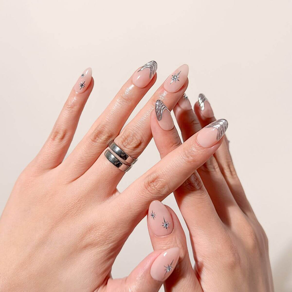 Model hands with a stacked silver ring displaying an elegant medium almond press-on manicure with sheer BB-cream pink base polish, futuristic silver chrome tips, and delicate star sparkles.