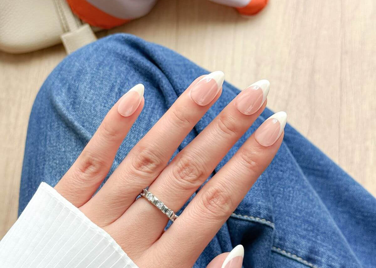 Eine Frau trägt künstliche Fingernägel zum Aufkleben in Mandelform im French-Nails-Design mit Chrome-Schimmer, kombiniert mit Jeans und weißer Bluse.