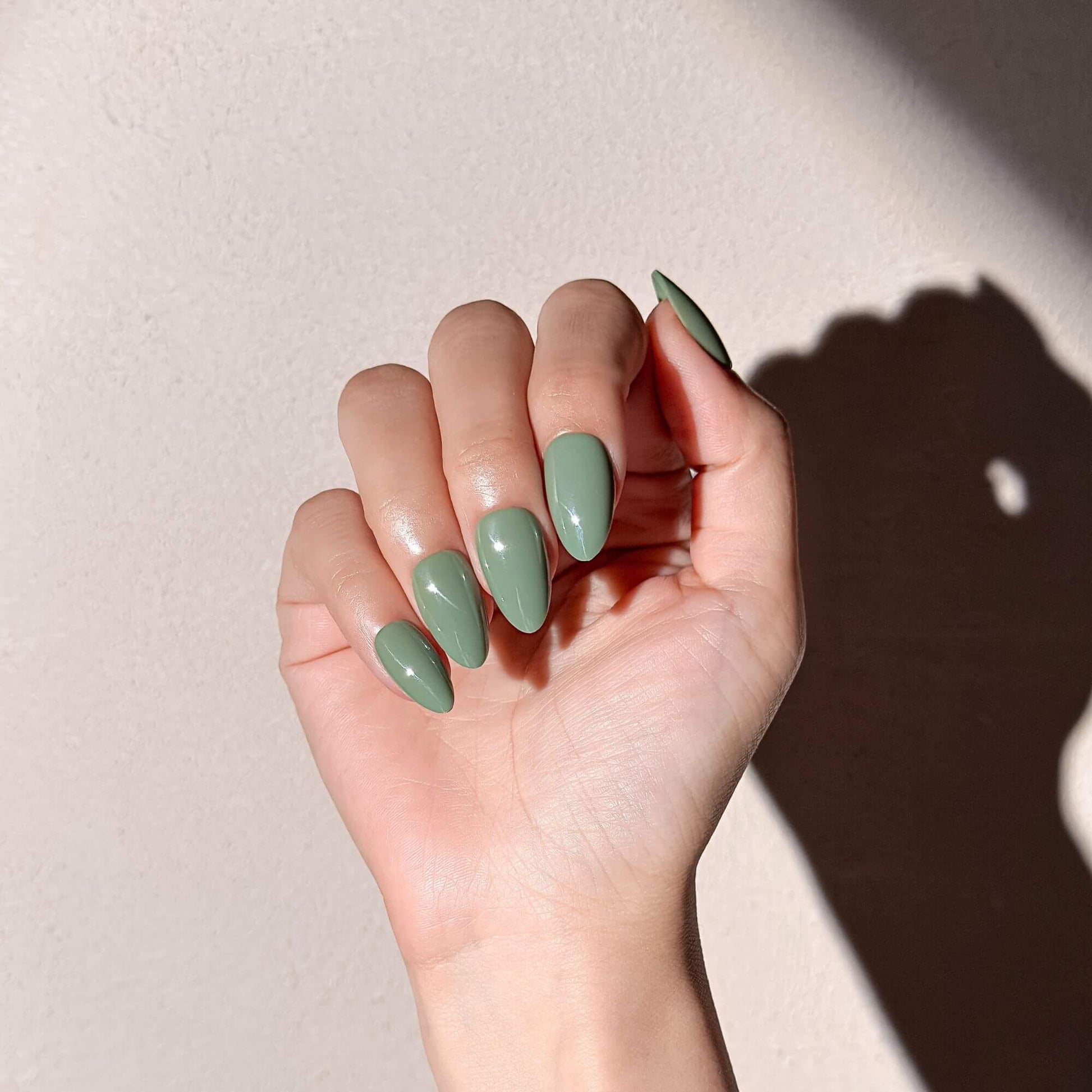 A hand showcasing a natural glossy green almond manicure under warm sunlight, highlighting the smooth finish and trendy earth-tone shade.