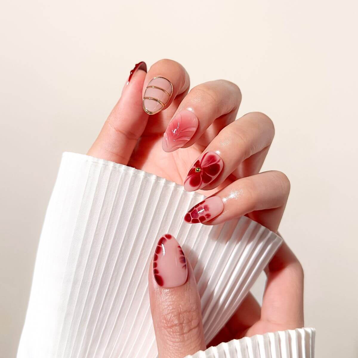 A close-up of a hand wearing glossy dark red almond-shaped glue-on nails with intricate 3D floral art and gold wire details.