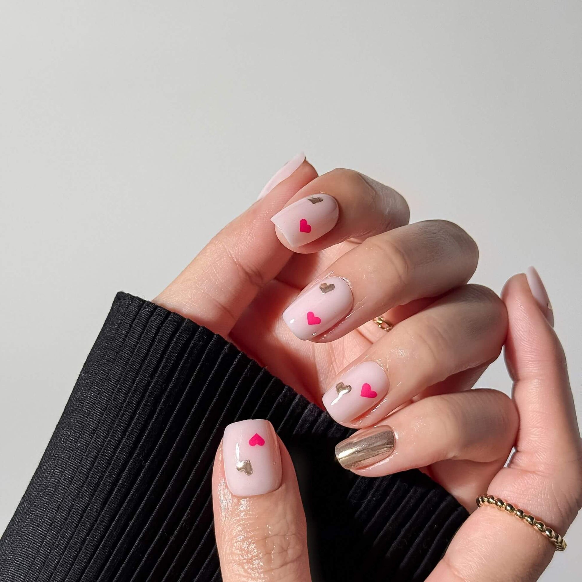 Close-up of a hand wearing short square milky pink nails with cute pink and silver metallic heart designs, styled with a gold ring and black sleeve.