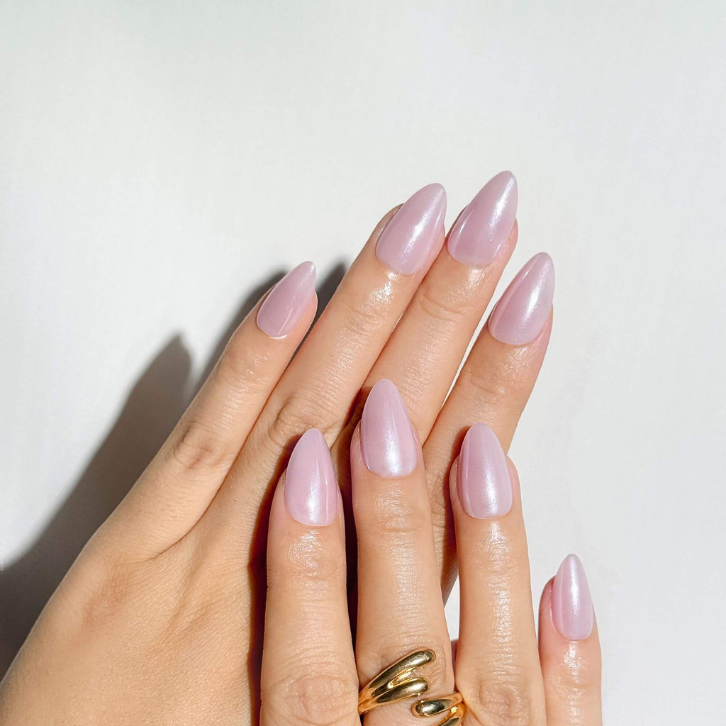 A detailed view of a soft glam manicure featuring shimmering pink almond-shaped press-on nails, showing the reflective pearl finish in natural light.
