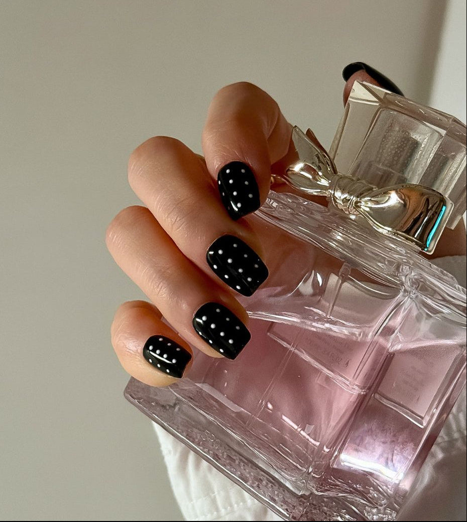 A hand wearing black press on nails with white polka dots in square short form holds a perfume bottle