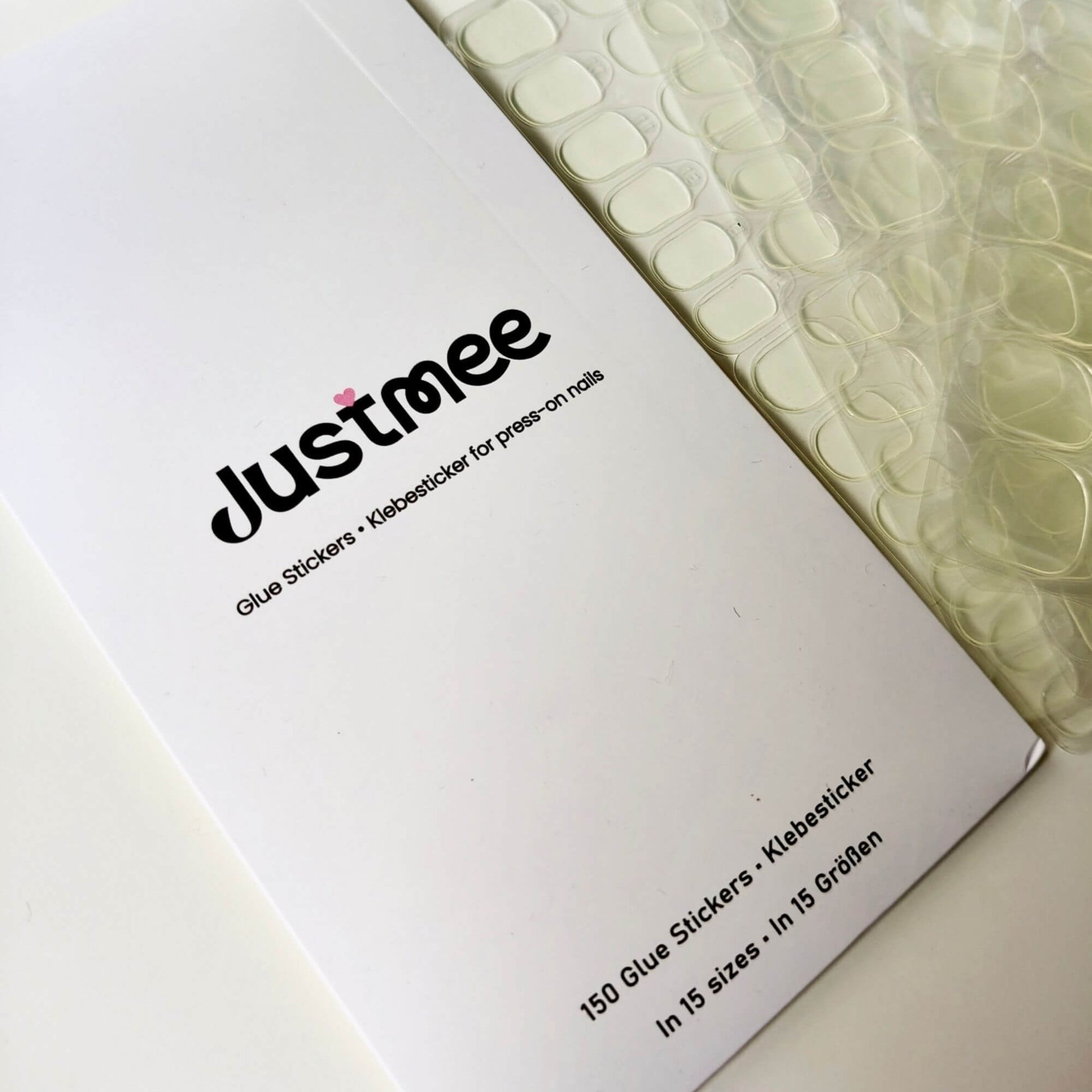 An overview of multiple sheets of transparent double-sided nail adhesive tabs next to their white branded envelope, showing the 150-piece refill set.