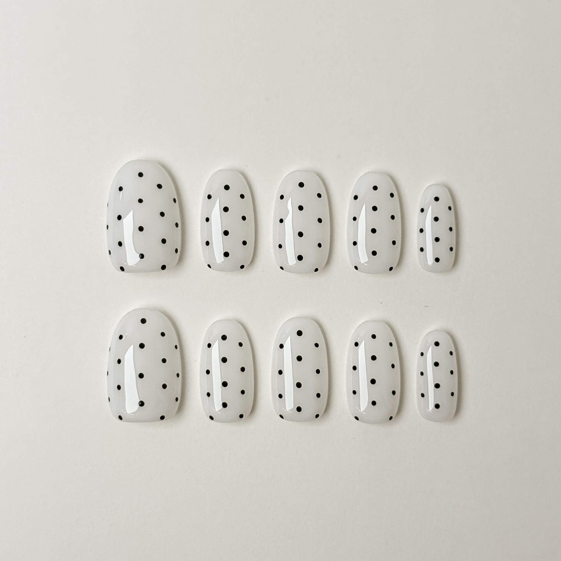 Eine Produktübersicht von 10 handgefertigten ovalen Press-on-Nägeln aus der Noir Dots Kollektion mit einer strahlend weißen Basis und minimalistischen schwarzen Punkten.