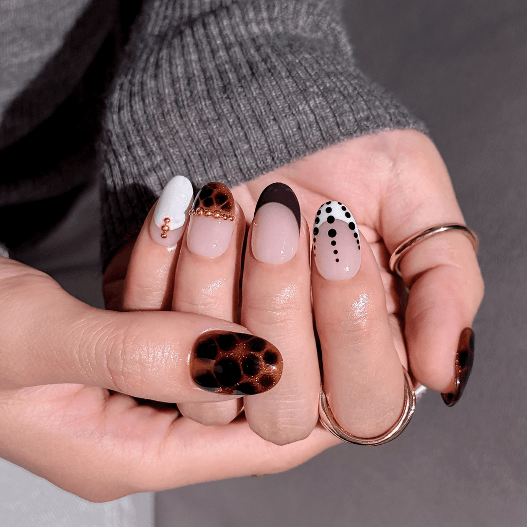 A close-up of a hand displaying a set of almond-shaped press-on nails with various designs, including leopard print, white with gold dots, black with white dots, and solid nude pink. The hand is wearing a gray ribbed sweater and gold rings against a neutral gray background.