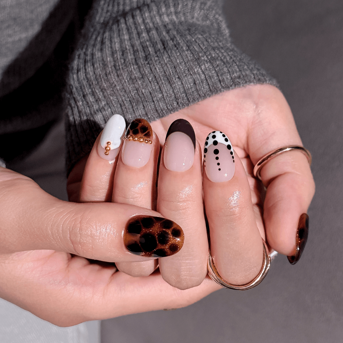 A close-up of a hand displaying a set of almond-shaped press-on nails with various designs, including leopard print, white with gold dots, black with white dots, and solid nude pink. The hand is wearing a gray ribbed sweater and gold rings against a neutral gray background.