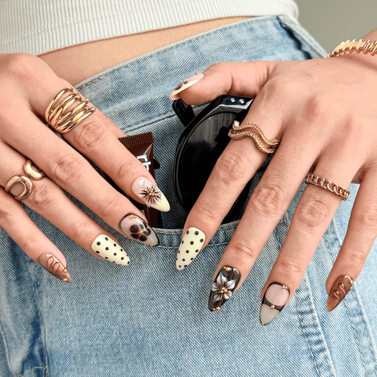 A close-up of a person's hands wearing multiple gold rings and long decorative almond-shaped nails in vintage black and nude with floral and dot designs resting on light blue denim jean.