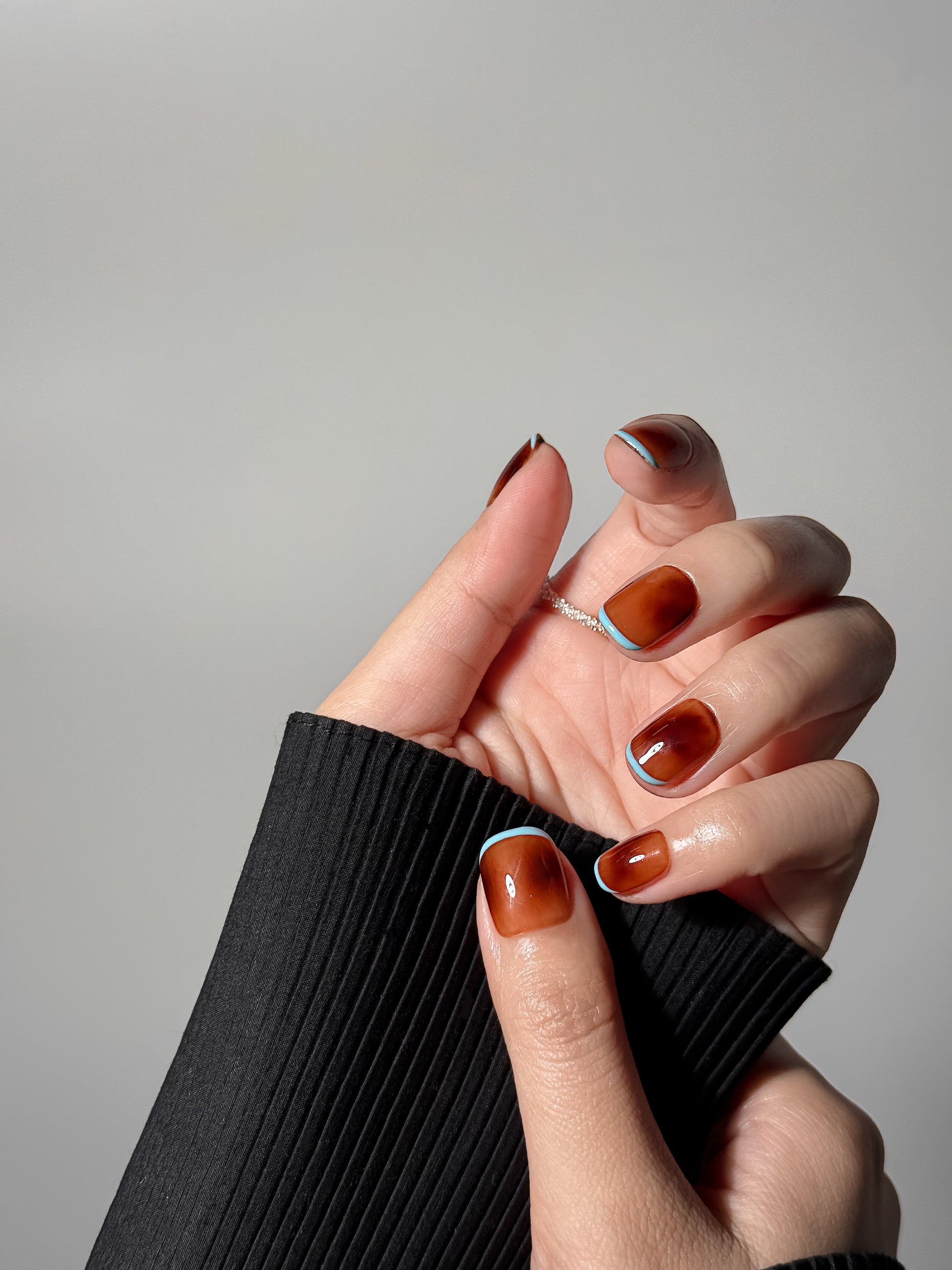 Close-up of a hand wearing short square amber-colored nails with light blue french tip detailing, paired with a thin silver ring and a black ribbed sleeve.