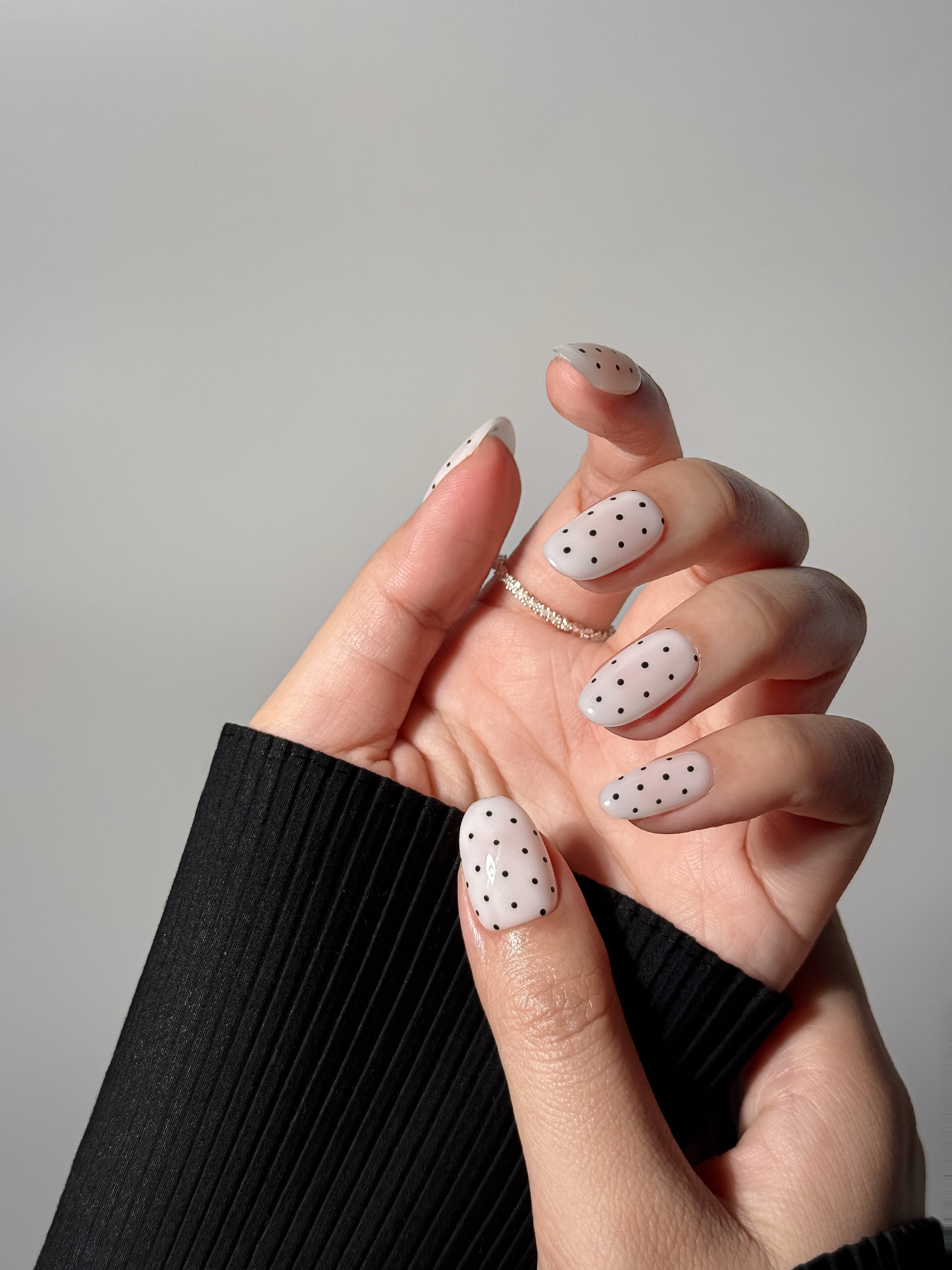 A hand wearing white nails with black polka dots, posed elegantly against a grey background with a black sweater sleeve.