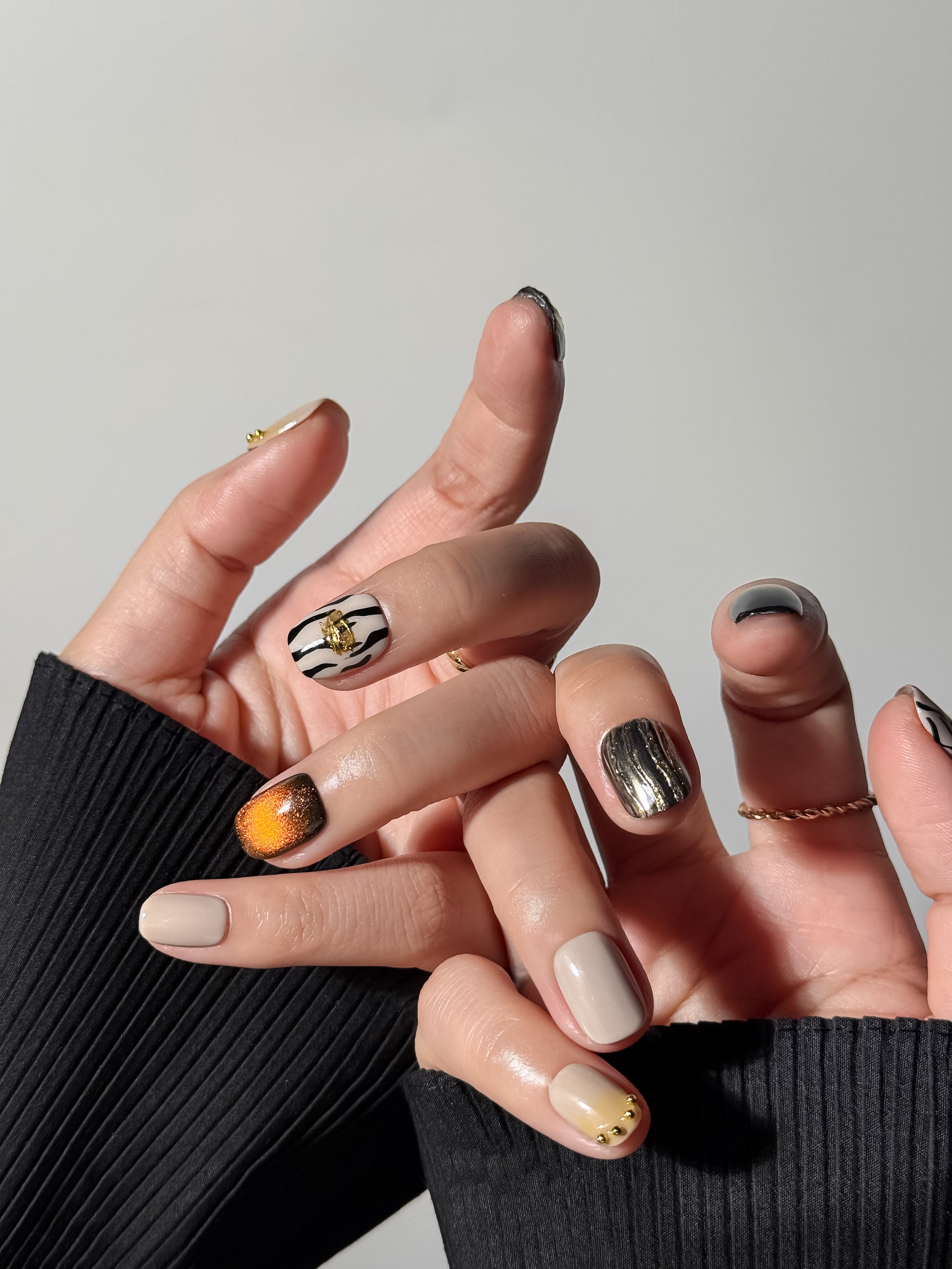 An artistic hand pose displaying a diverse nail art design including textured silver metallic ridges, zebra animal patterns, and nude nails embellished with tiny gold studs along the cuticle.