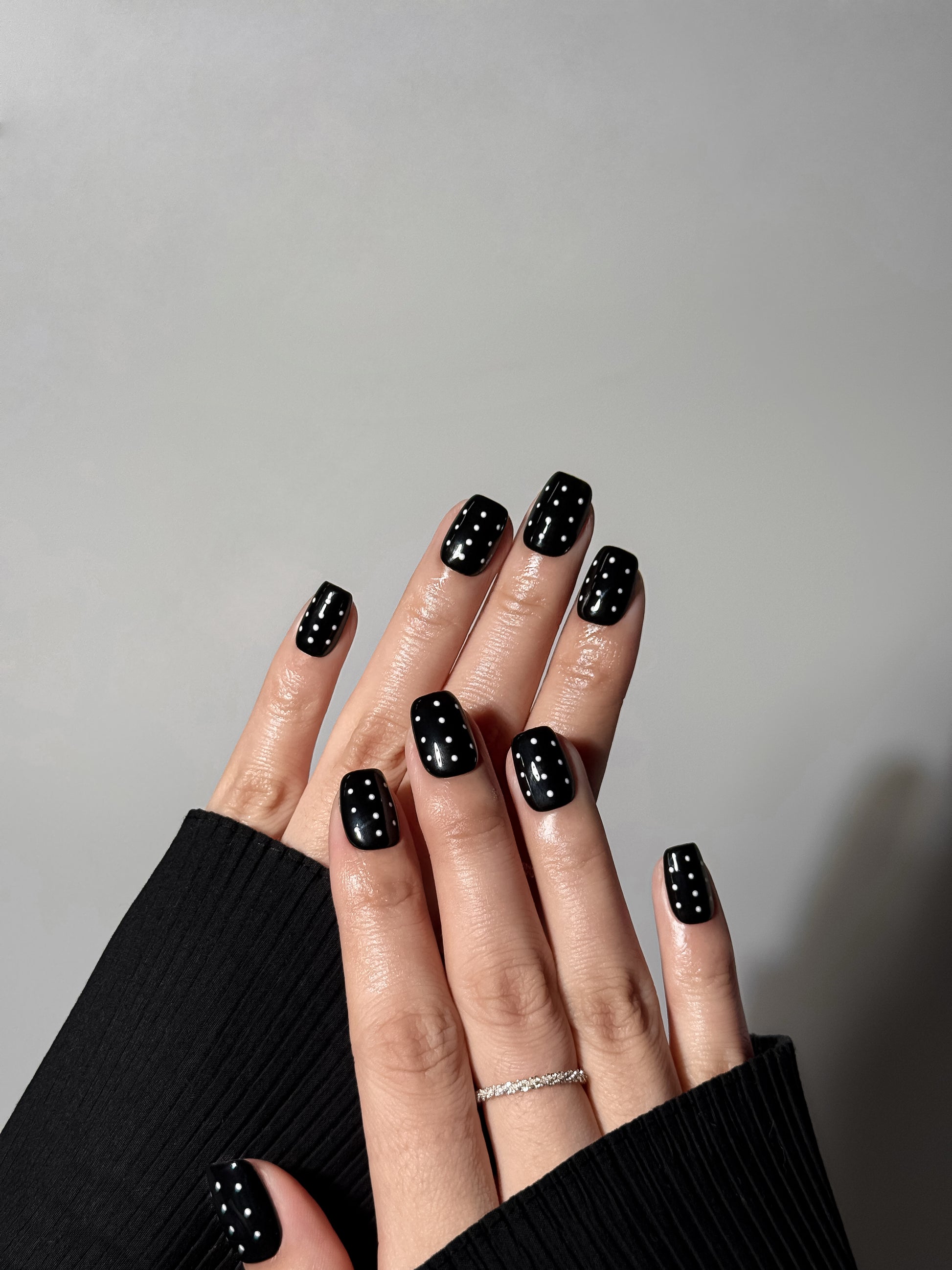 Both hands displaying a bold black manicure with vertical white dot accents, styled with a black ribbed sweater and silver ring