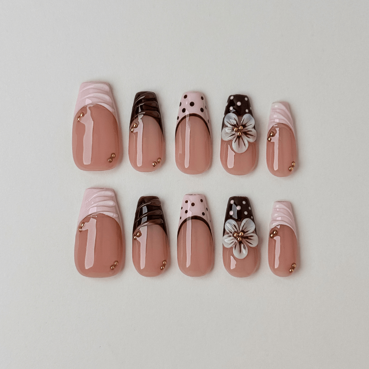 A set of ten false press-on nails arranged in two rows on a light gray background. The nails feature a glossy nude pink base with various decorative designs including white satin-like tips, dark brown polka dots, white cherry blossom flowers, and small gold metallic accents.