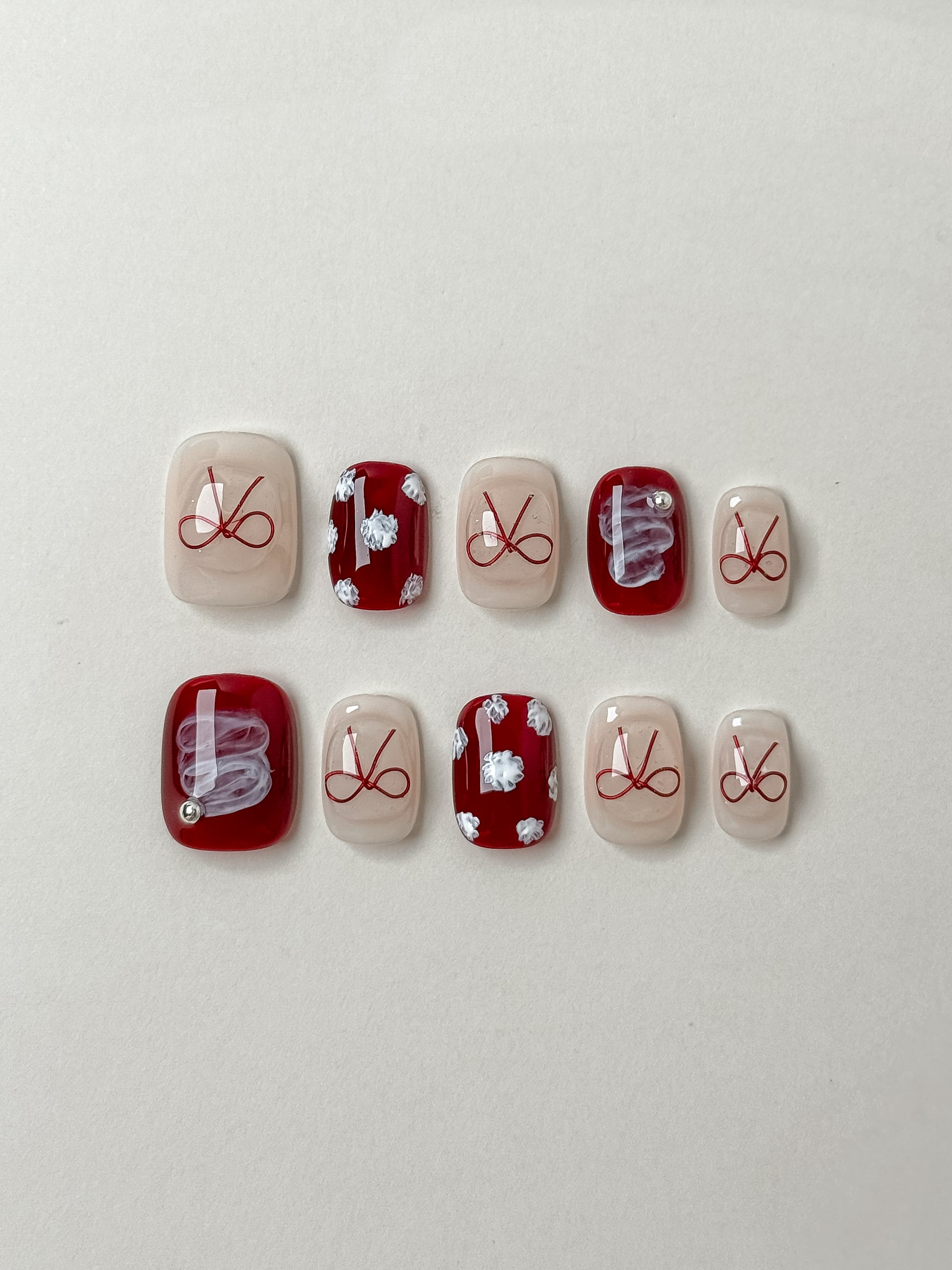 flat-lay display of coquette press-on nails with red ribbon bows, white floral patterns, and swirl accents on short square shapes.