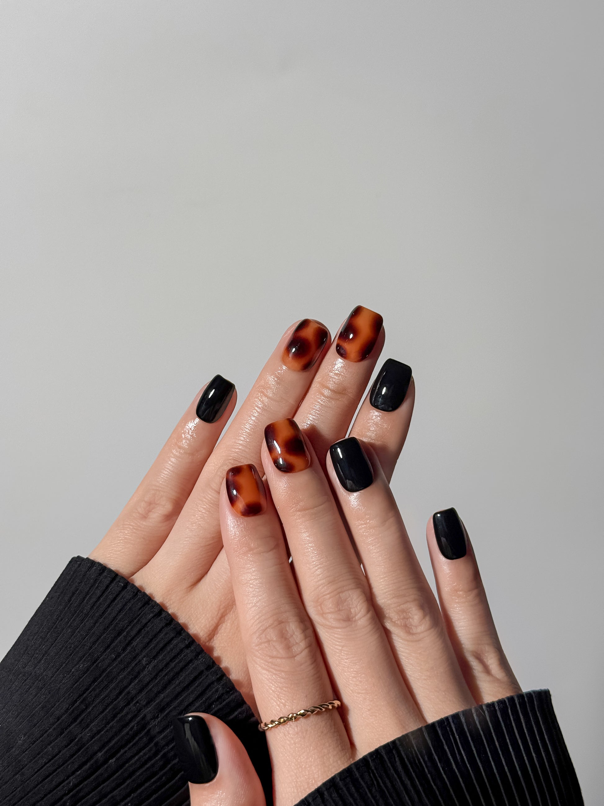 Two hands showcasing a bold mix of solid black and tortoiseshell print press-on nails, styled with a gold twisted ring.