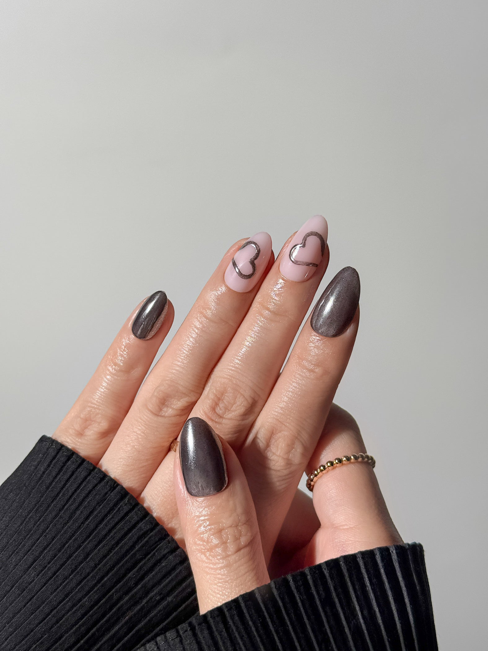 Close-up of a metallic heart-themed manicure with graphite chrome accents styled with gold rings.