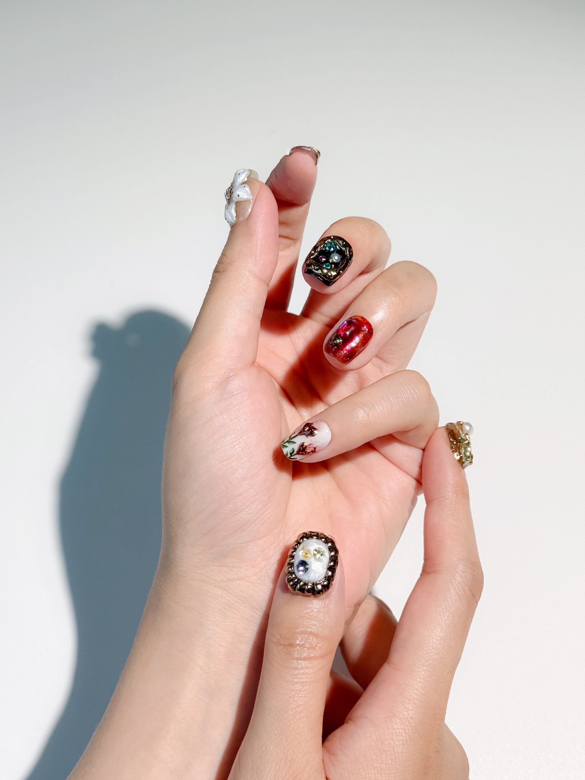 A hand wearing ornate, jewelry-inspired nails including a white thumb nail with a gold textured frame and gems, a ruby red glitter nail, and a 3D white flower charm with pearl centers.